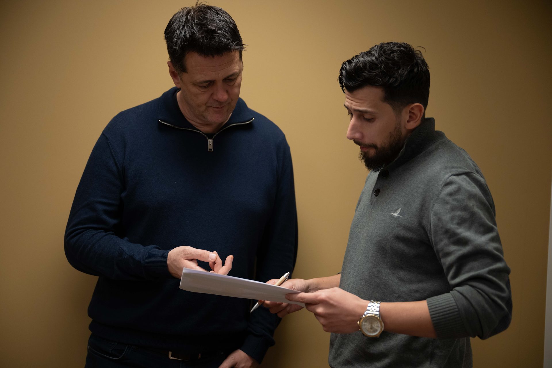 Two men in sweaters reviewing a document together, standing against a tan wall.