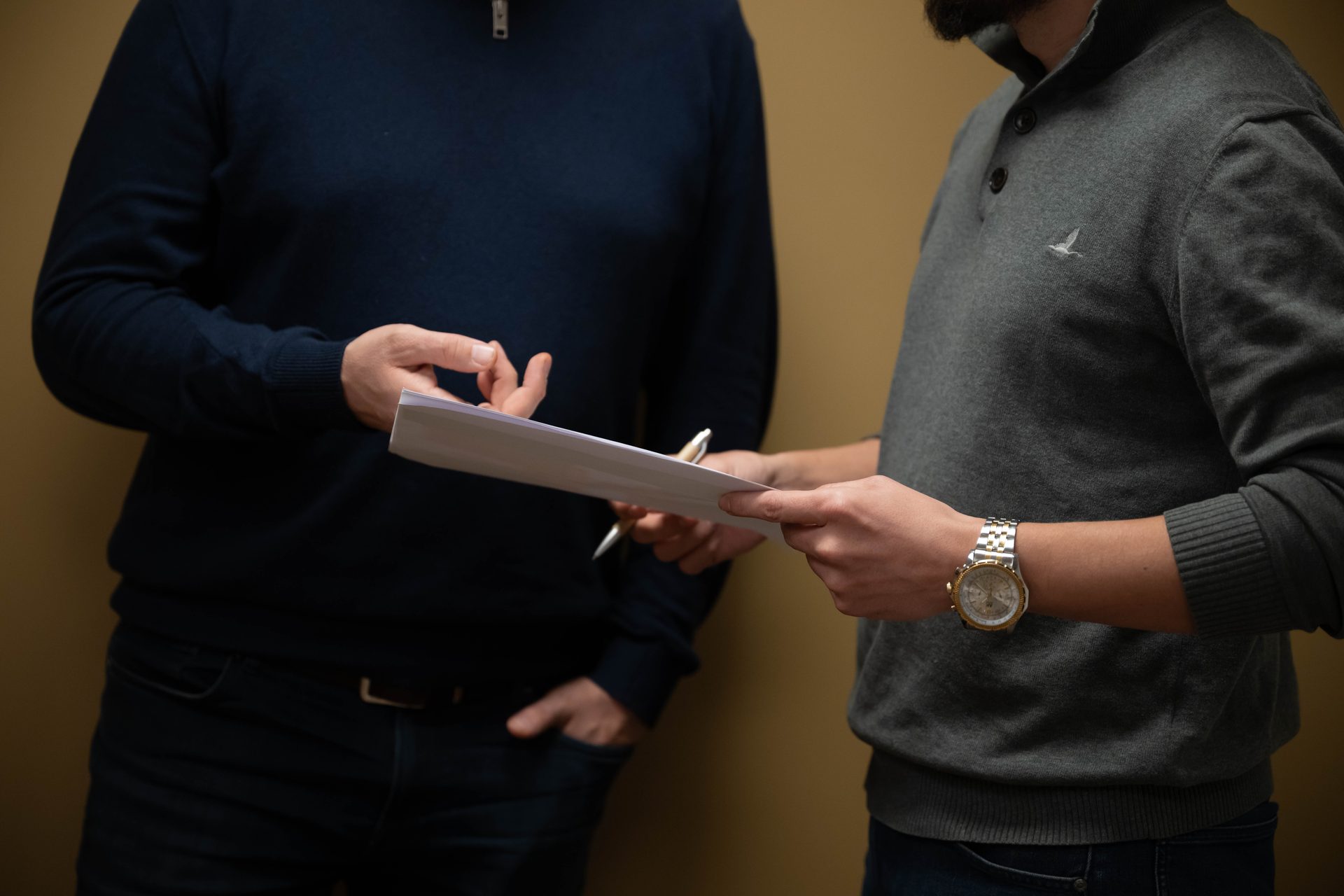 Two people are shown from the chest down, exchanging or reviewing documents and a pen.