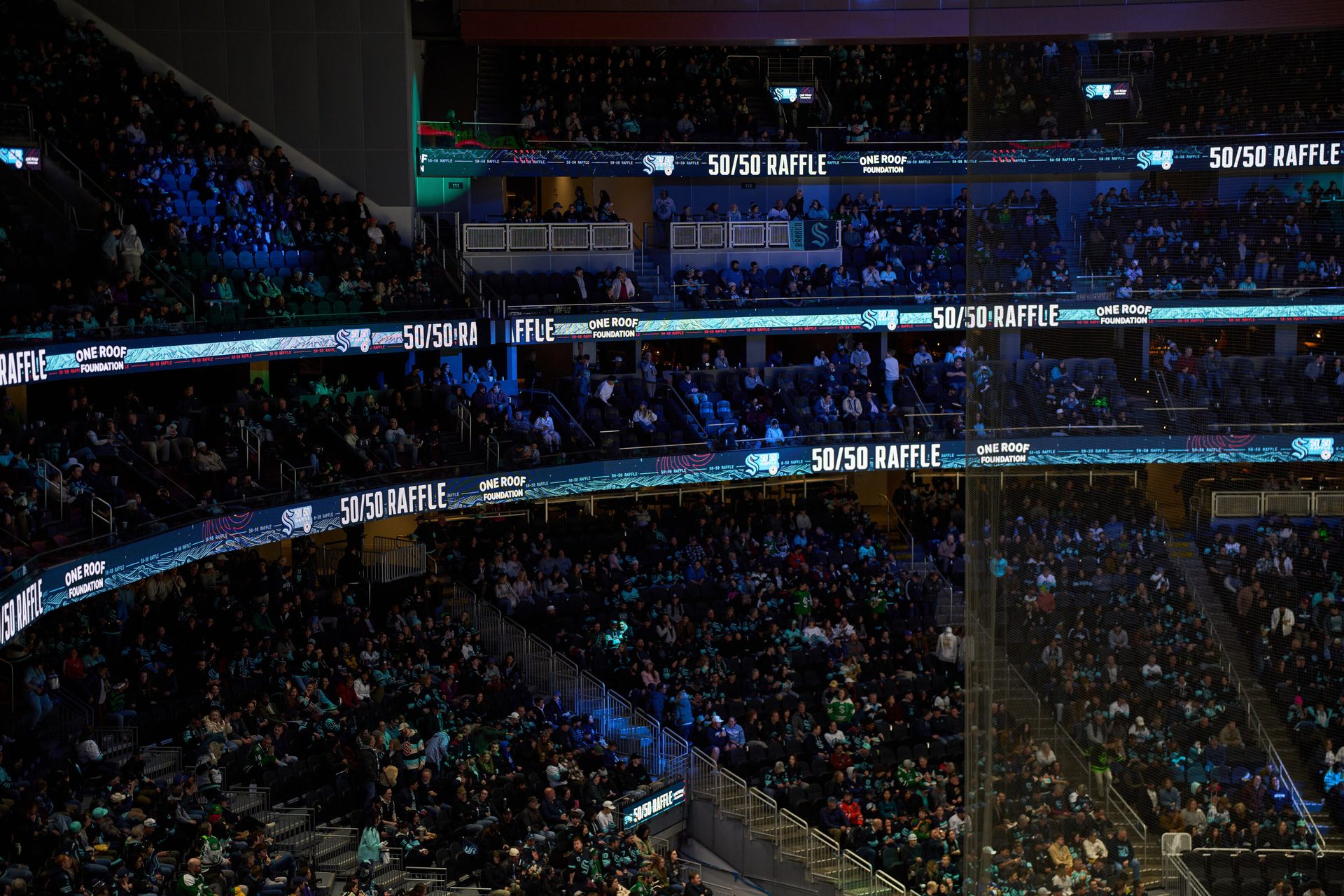 Indoor arena crowd with multiple LED signs displaying "50/50 RAFFLE" and "ONE ROOF FOUNDATION."