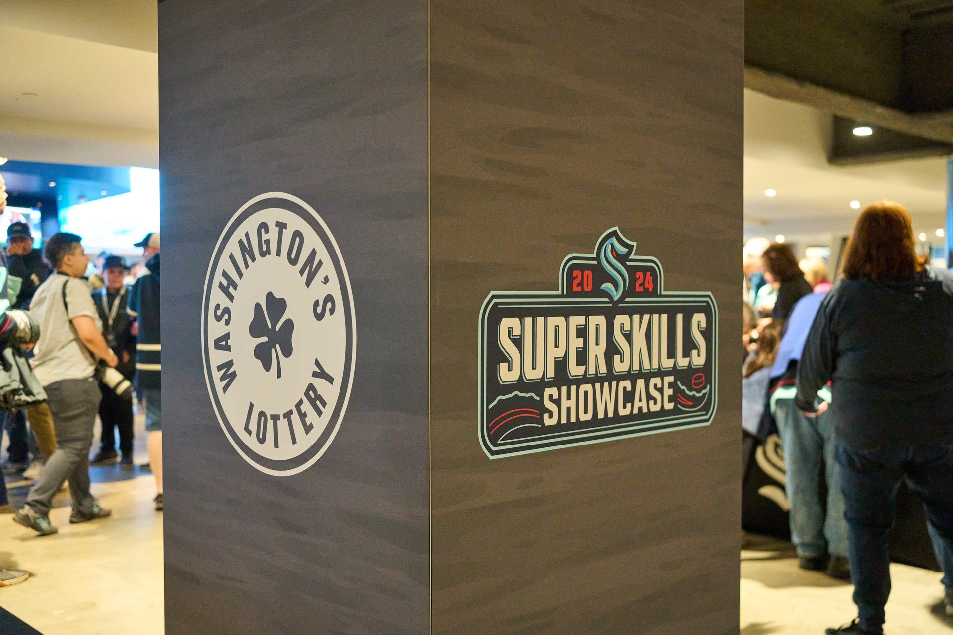 Two signs: Washington's Lottery and Seattle Kraken 2024 Super Skills Showcase with people.