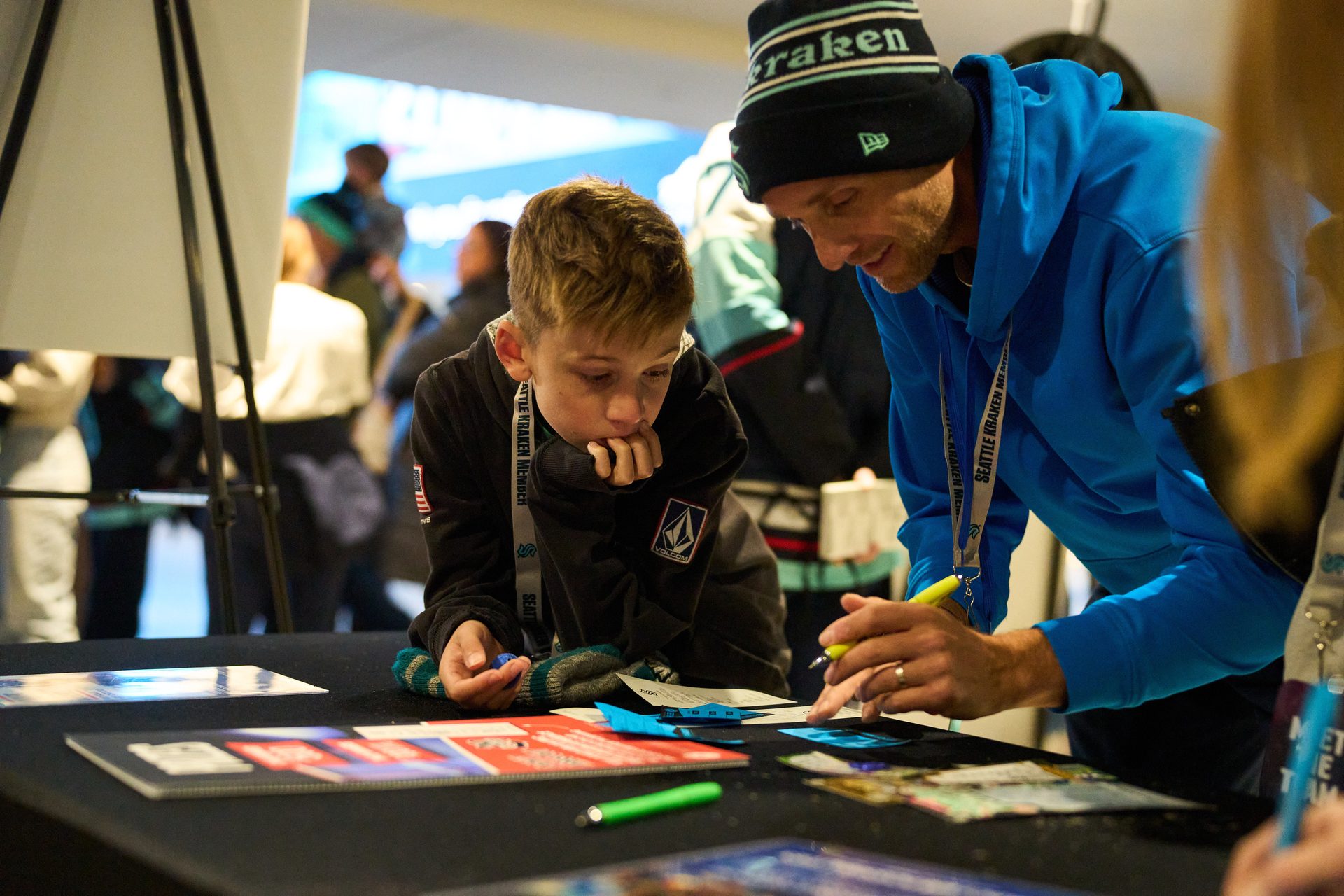A man in a Kraken beanie and boy examine items on a table, both wearing Seattle Kraken lanyards.