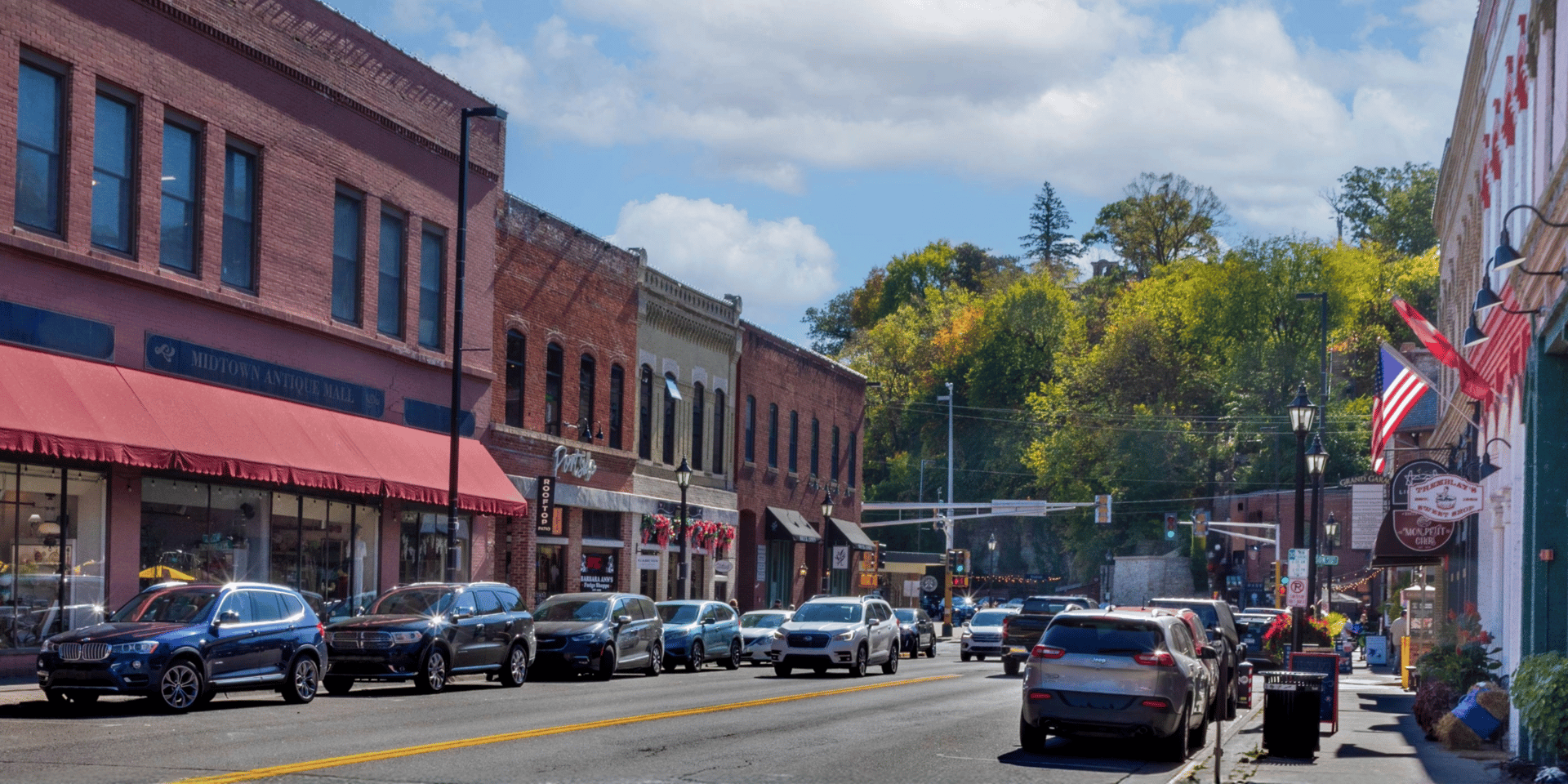 A sunny street scene in a small town with historic brick buildings, parked cars, and lush trees.