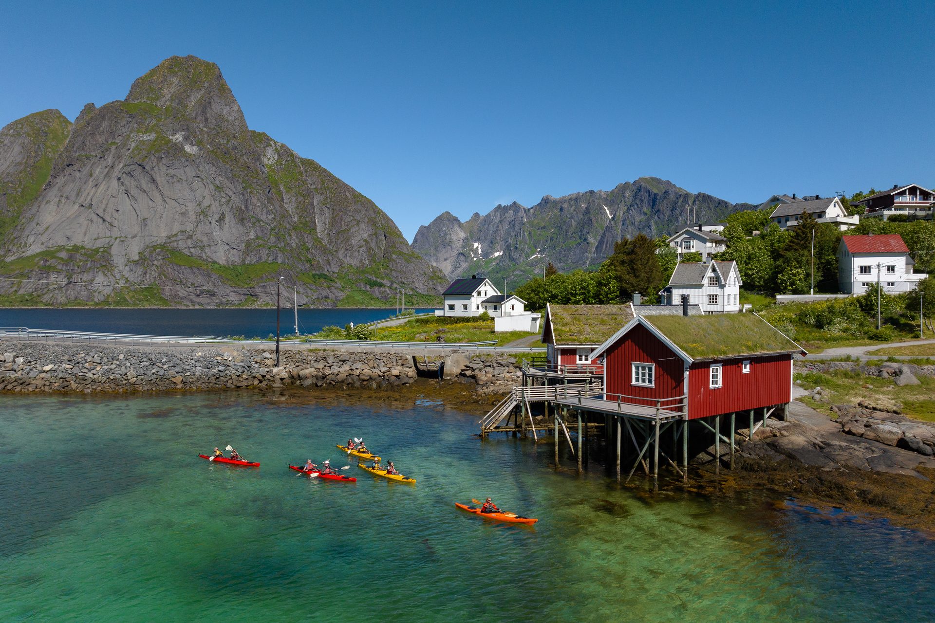 Kayakers on clear water near red stilt houses and mountains under blue sky in scenic village.