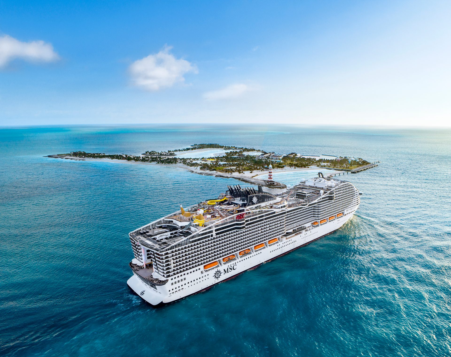 Aerial view of a large MSC cruise ship sailing near a pristine tropical island with a lighthouse.
