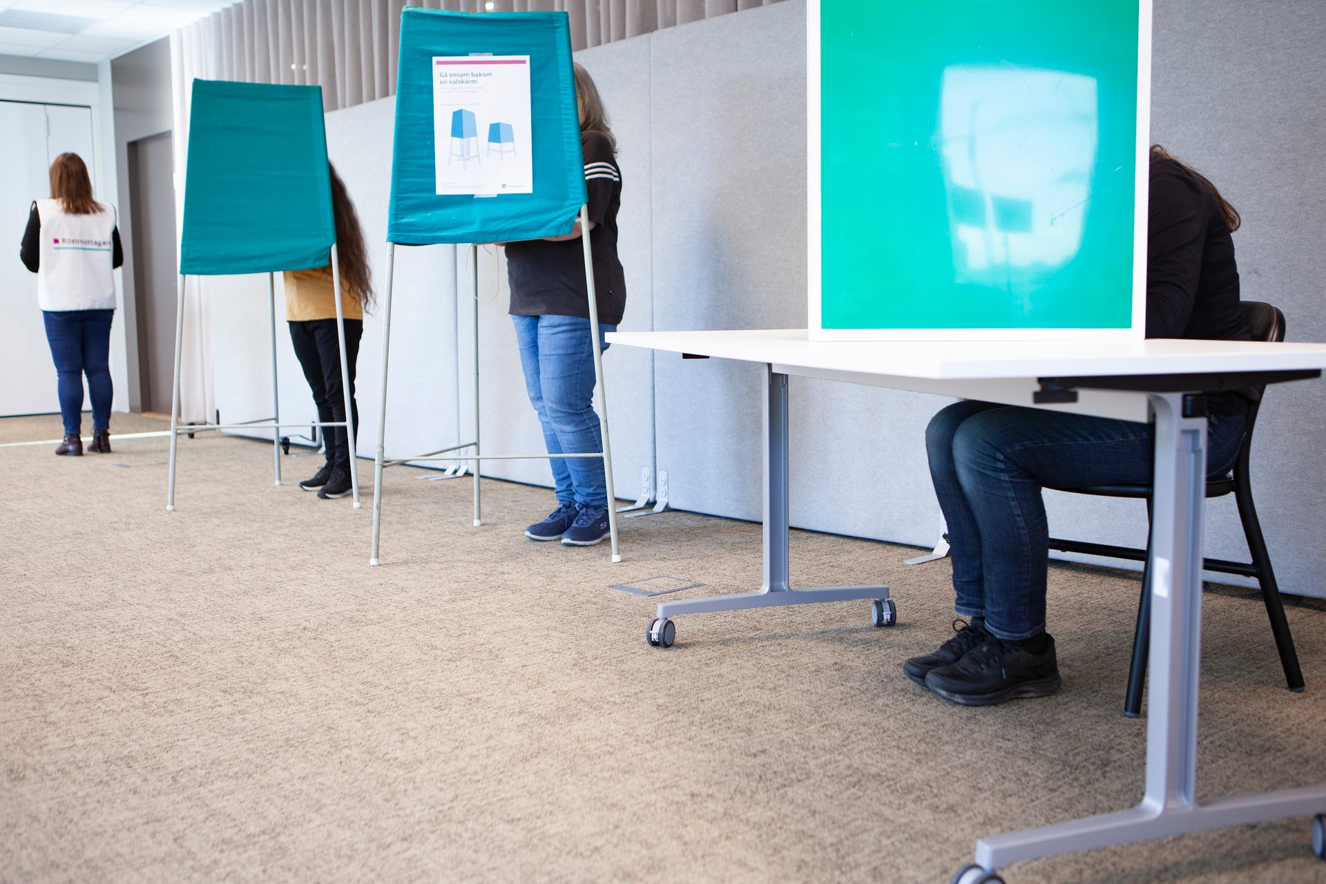People standing and sitting at green voting booths in a room.