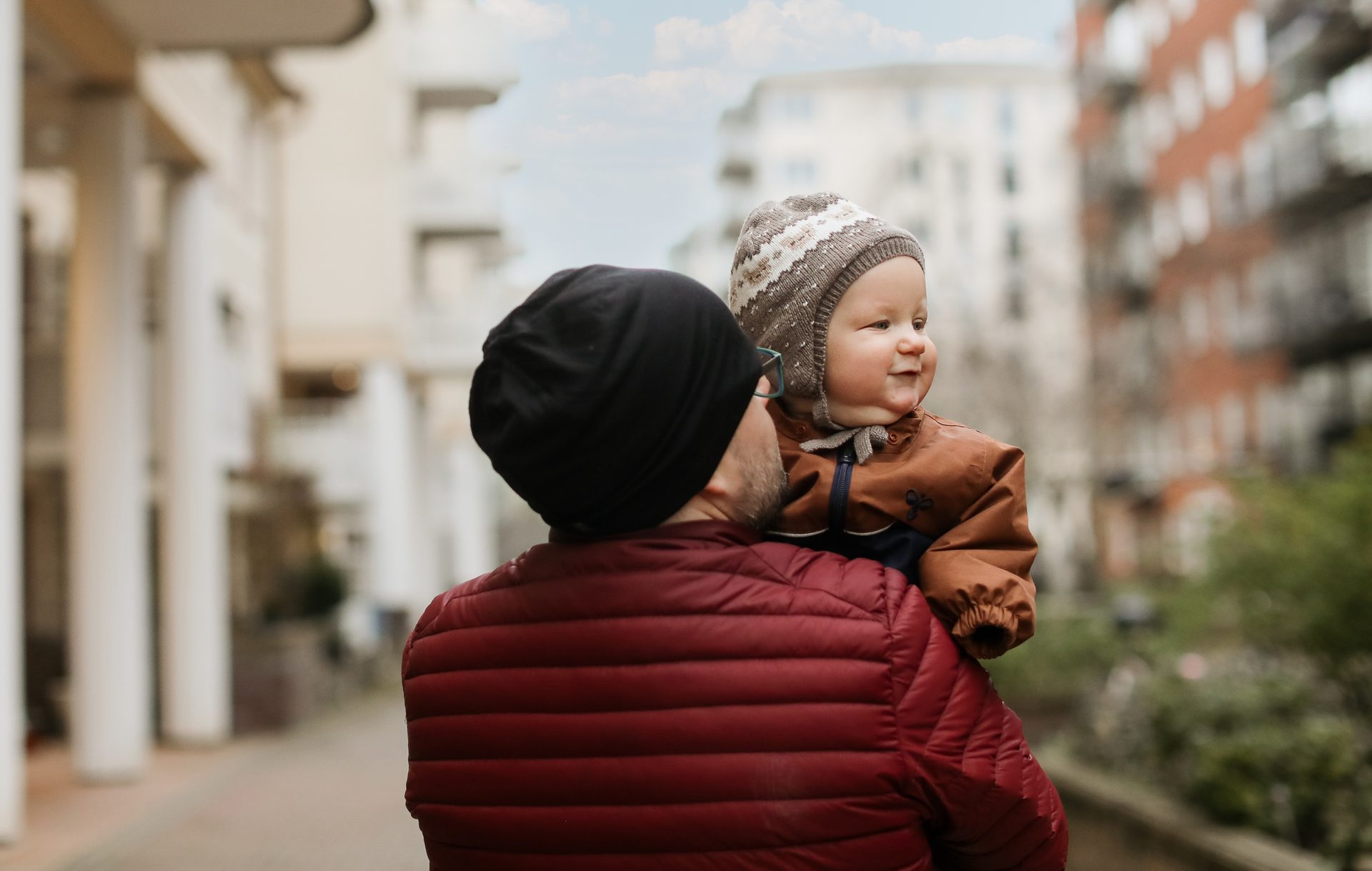 An adult carries a smiling baby, wearing a patterned hat, on their shoulders outdoors.