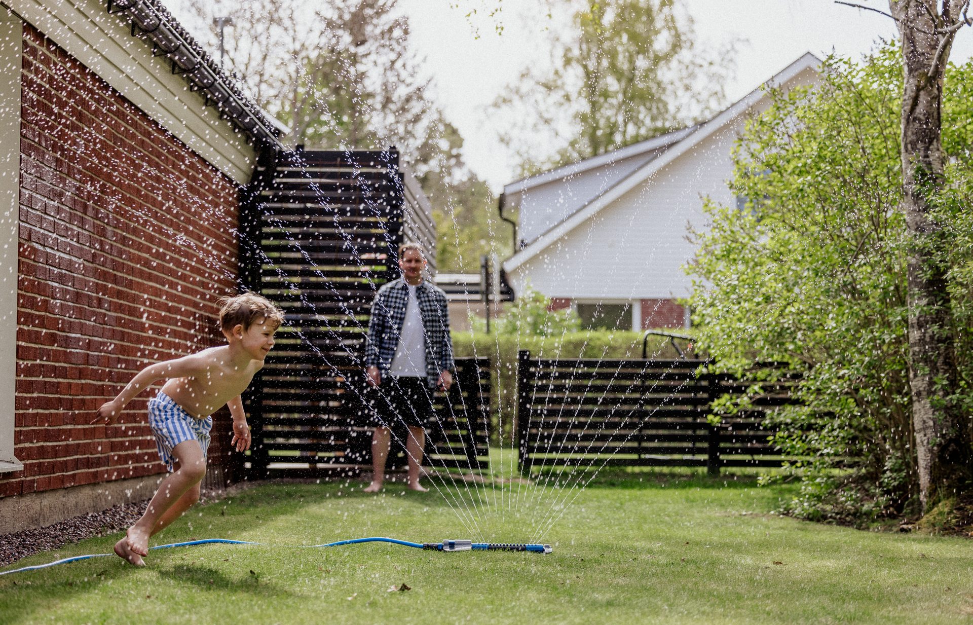 A boy plays in a sprinkler in a green yard, with a man standing in the background.