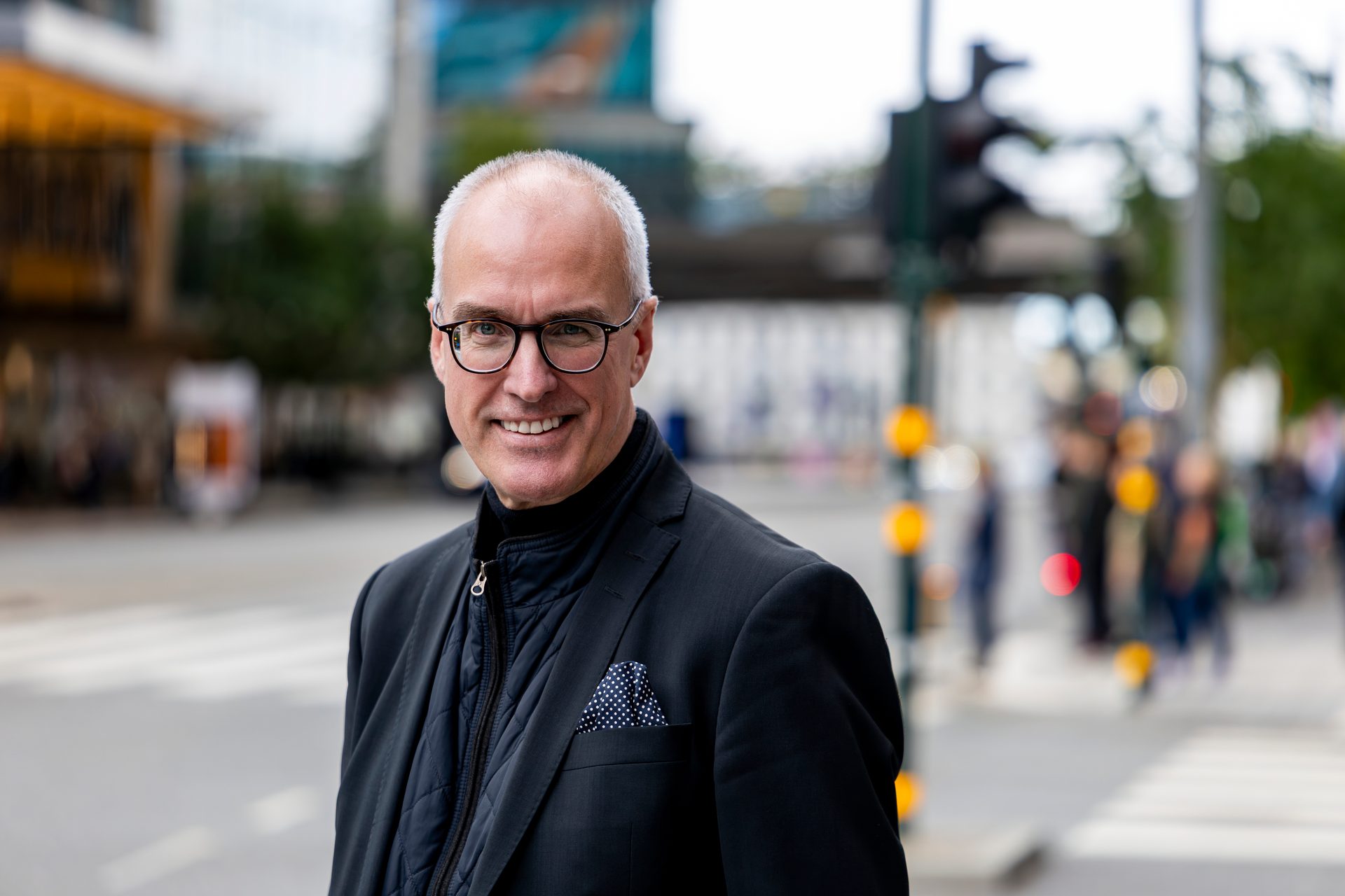 Smiling man in glasses and suit jacket on a blurred city street.
