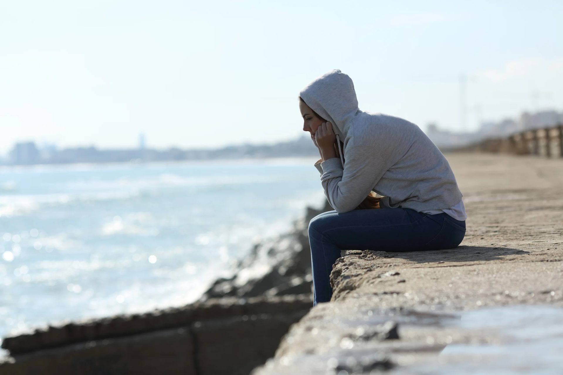 A young woman in a hoodie sits sadly by the ocean, chin in hands.