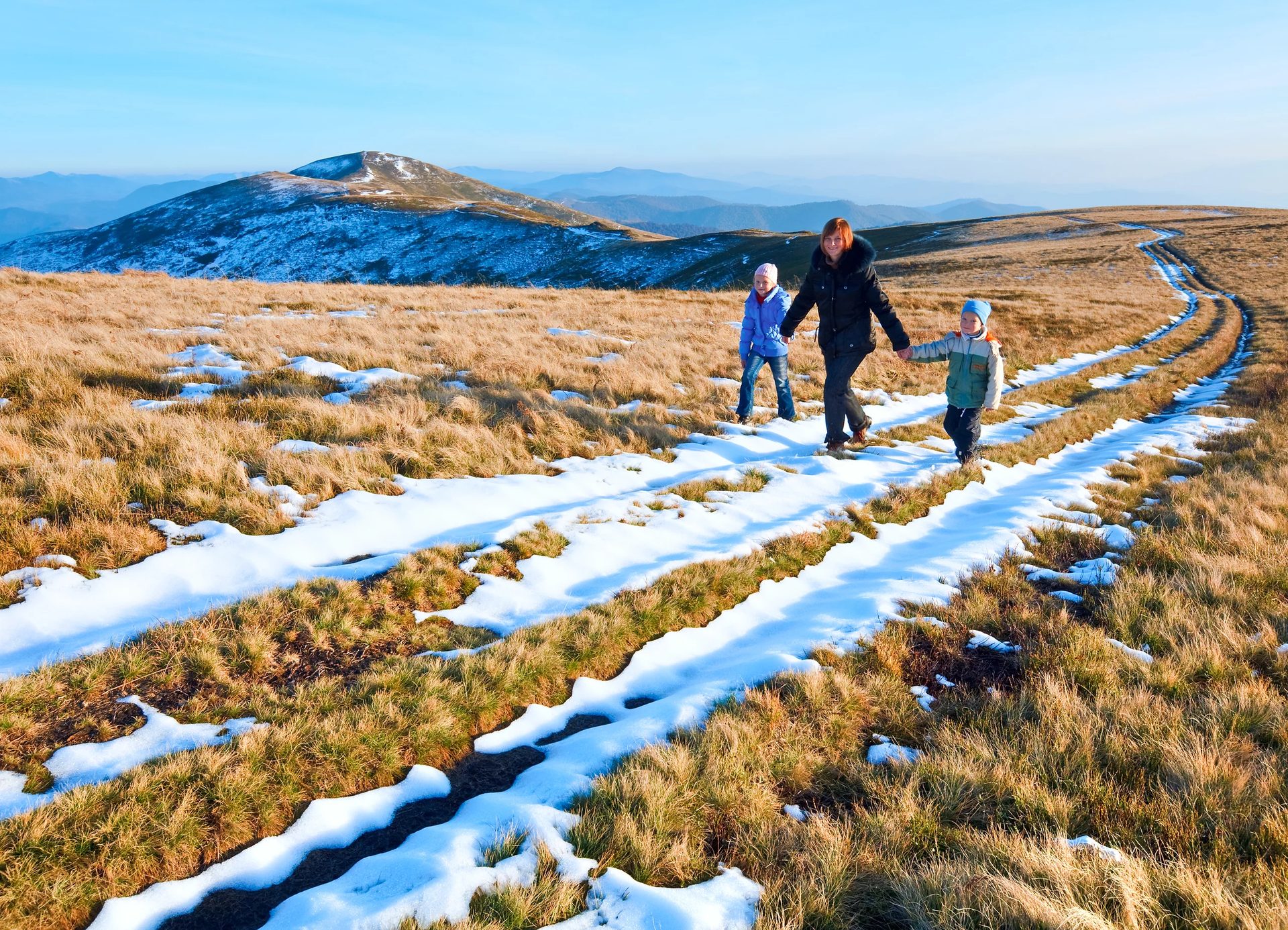 Family walks a snowy, grassy mountain path with distant peaks under a blue sky.