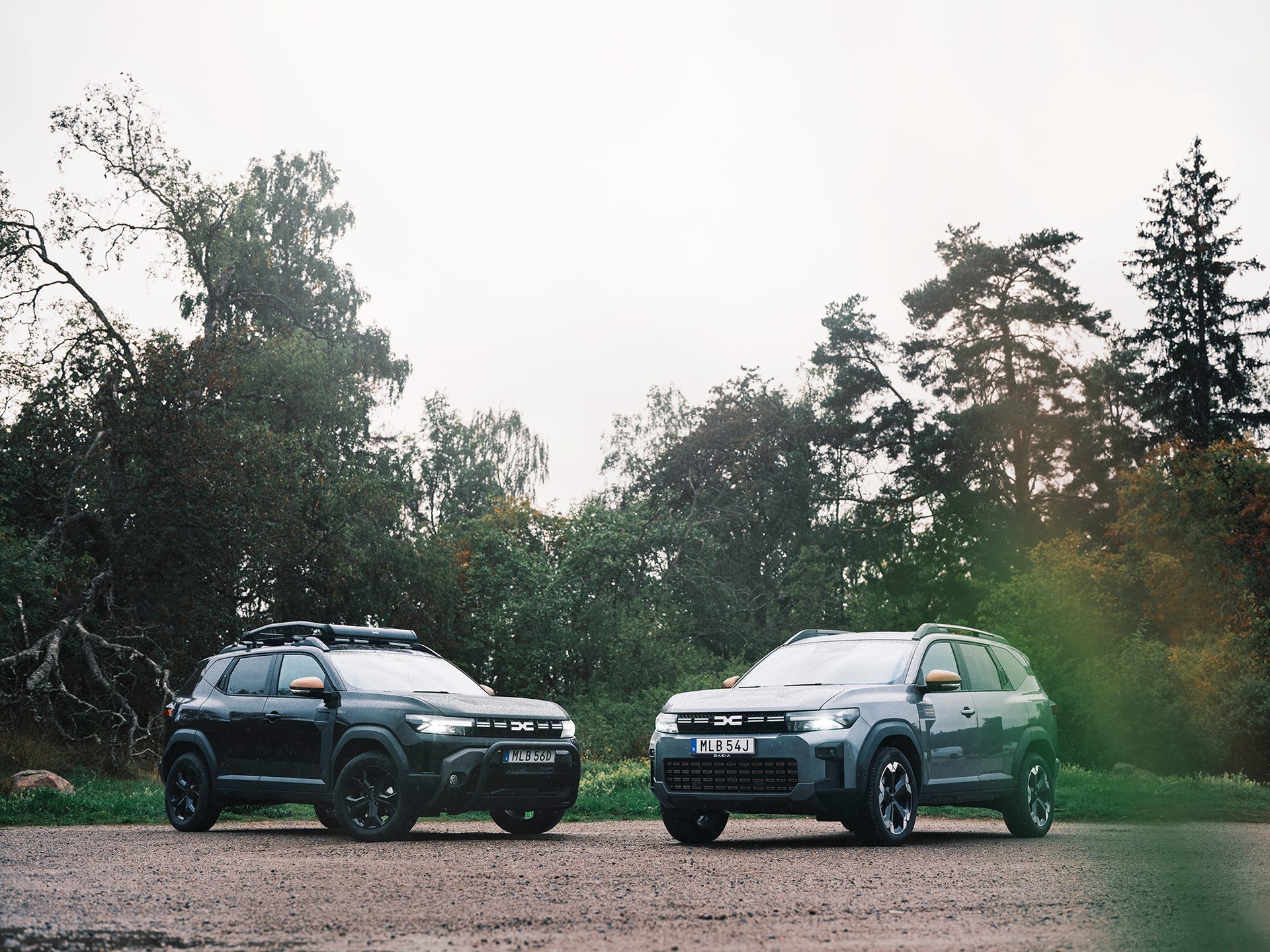 Two Dacia Duster SUVs, one dark and one gray, parked on a dirt road in front of trees.