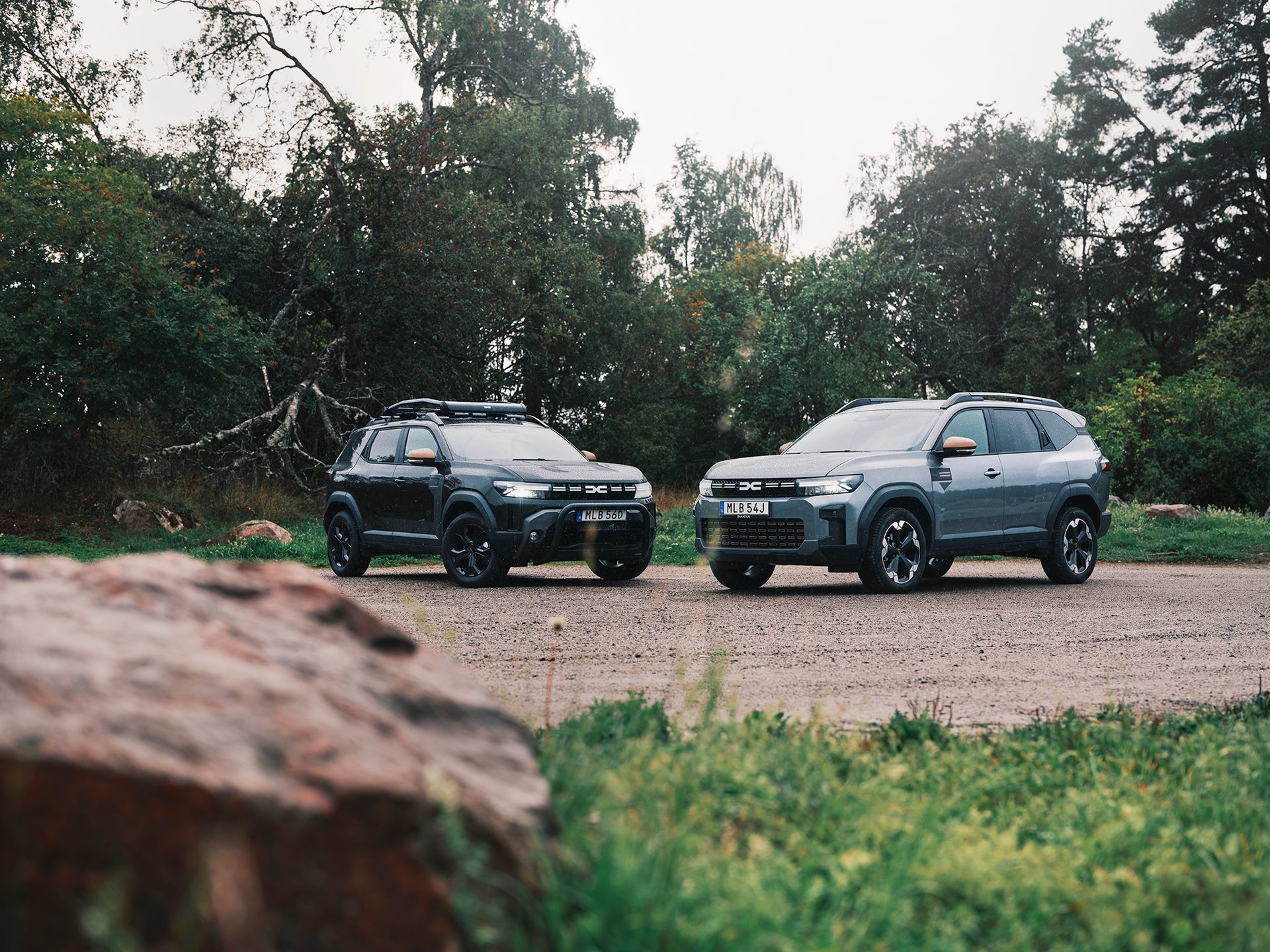 Two Dacia Duster SUVs, one black and one grey, parked on a dirt road amidst trees on a cloudy day.