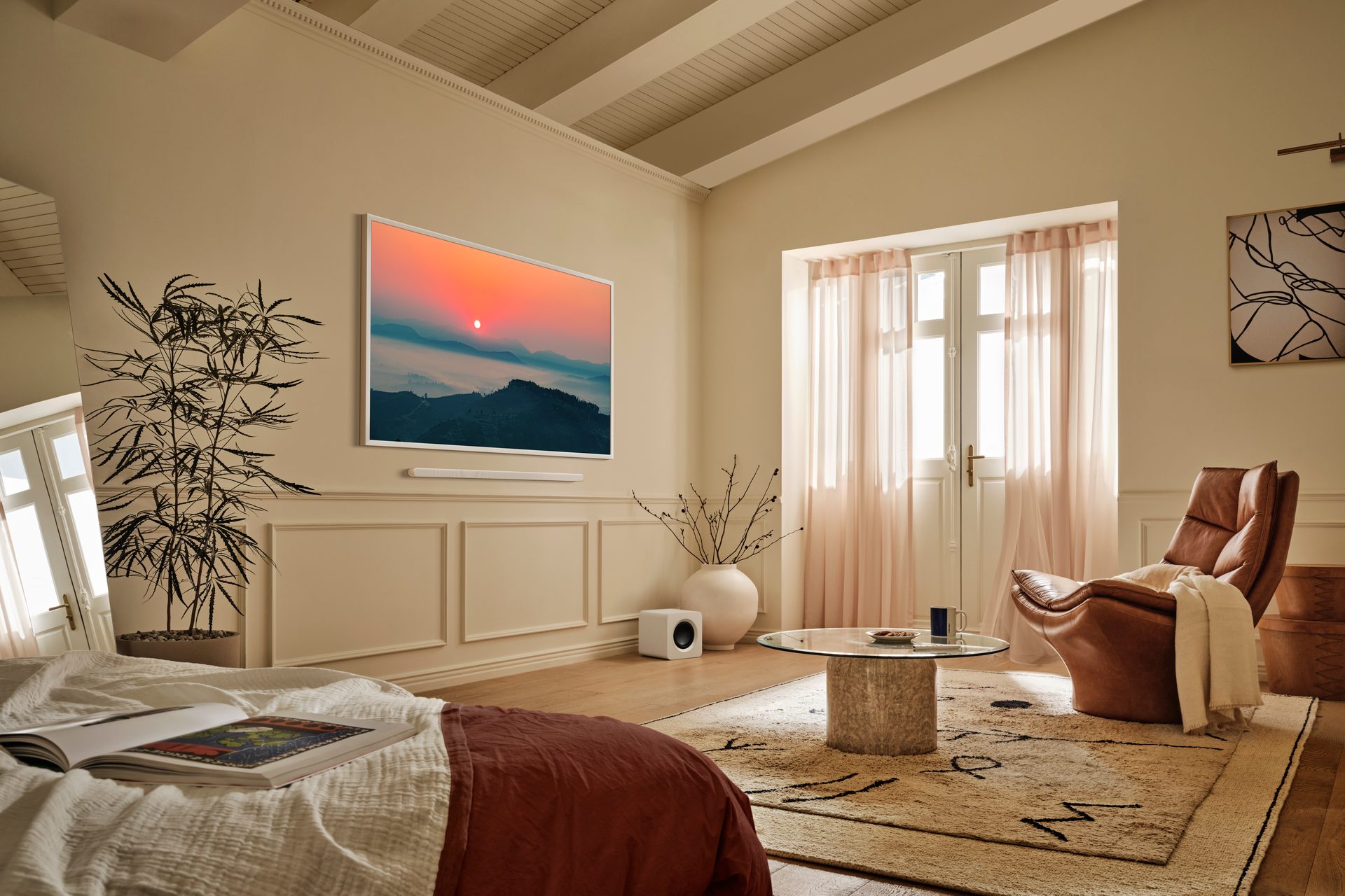 Bright, neutral room with bed, TV showing sunset, brown leather armchair, and large window.
