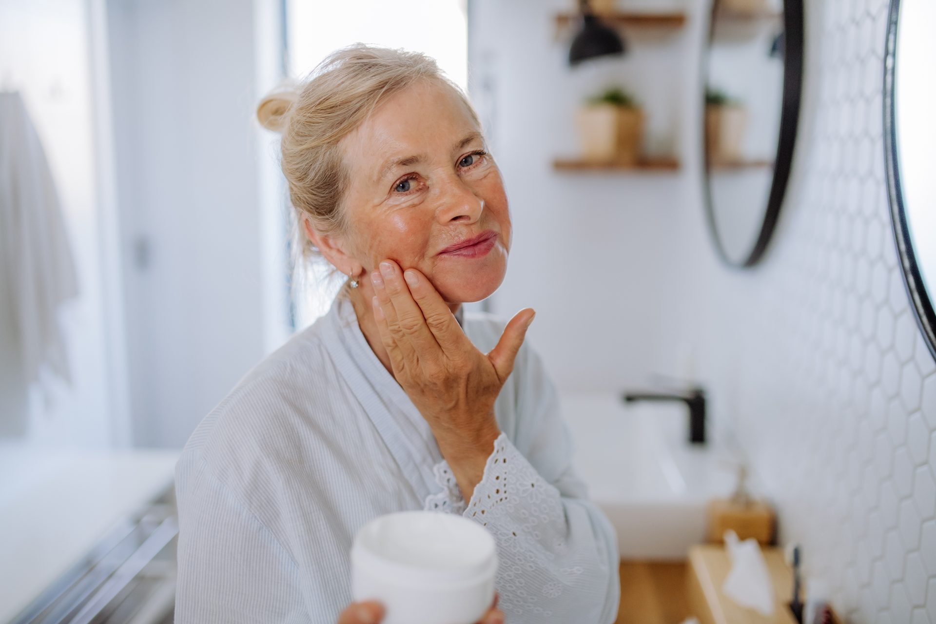 Older woman in a bathroom applying face cream to her face, looking at the camera.
