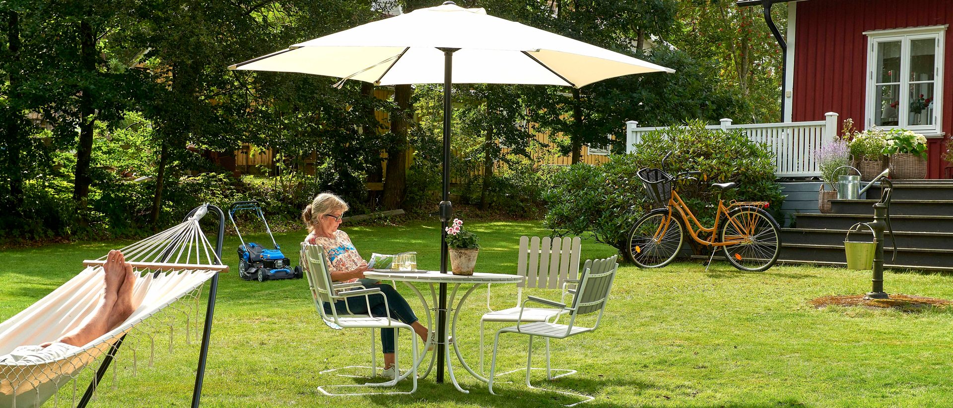 A woman reading under an umbrella, a hammock, and a red house in a sunny garden.