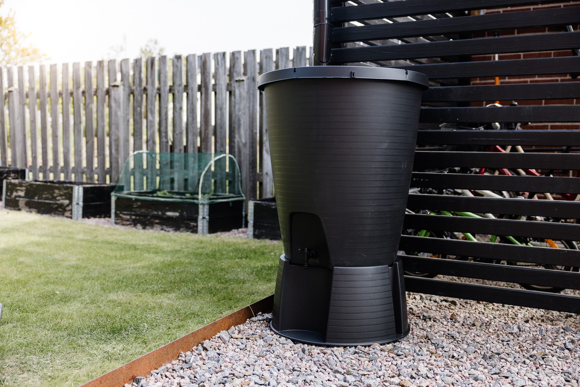 Black rain barrel on gravel next to a dark fence in a backyard.