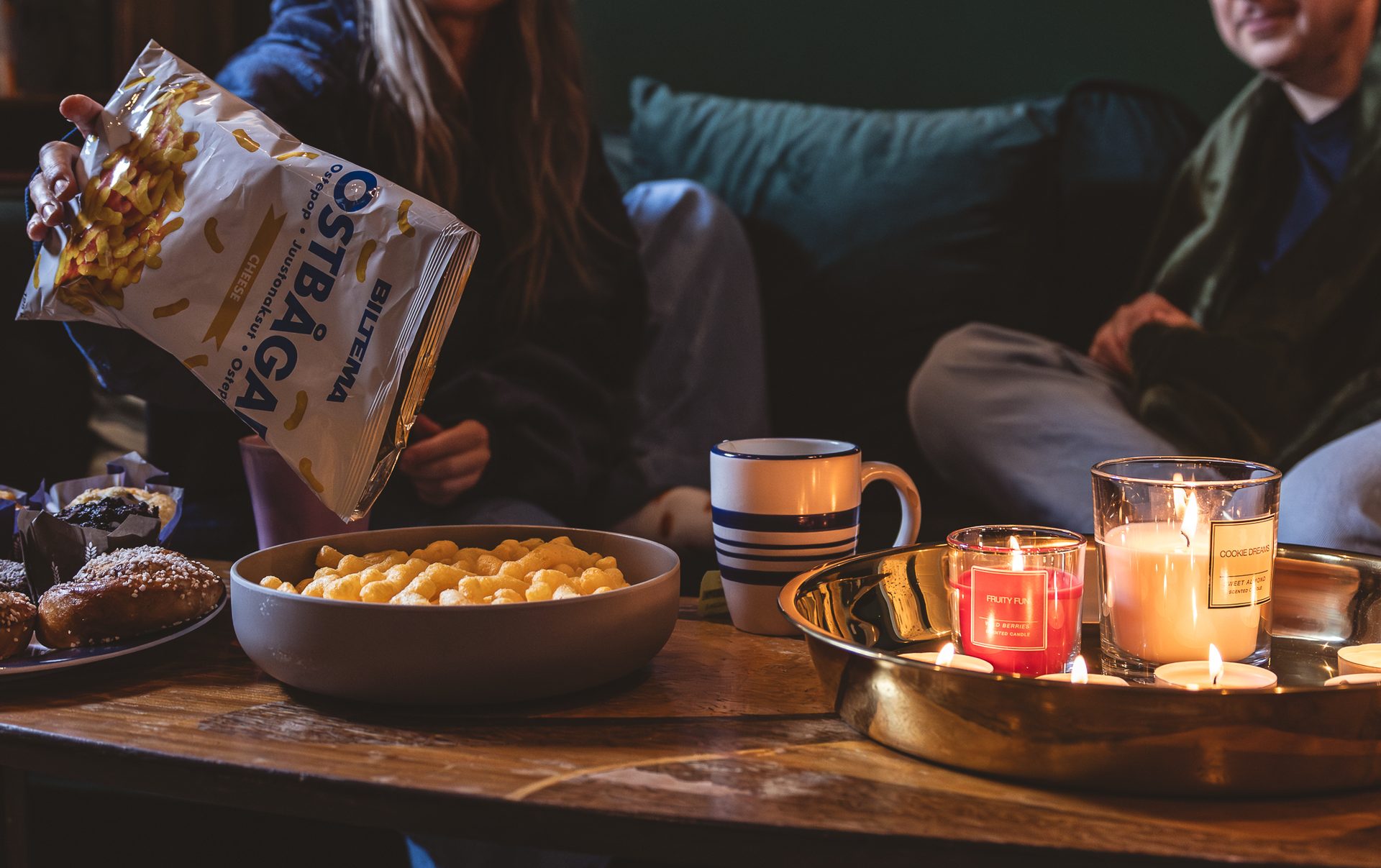 Person pouring cheese puffs into a bowl, with muffins, a mug, and lit candles on a coffee table.