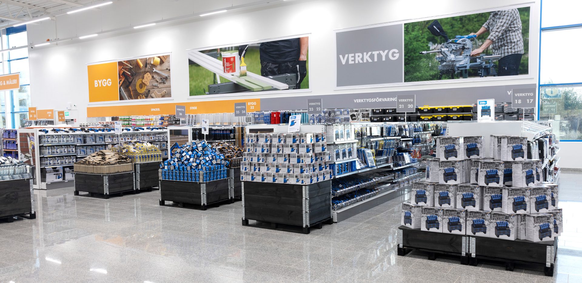 Brightly lit hardware store interior with shelves and bins of tools, building materials, and paints.