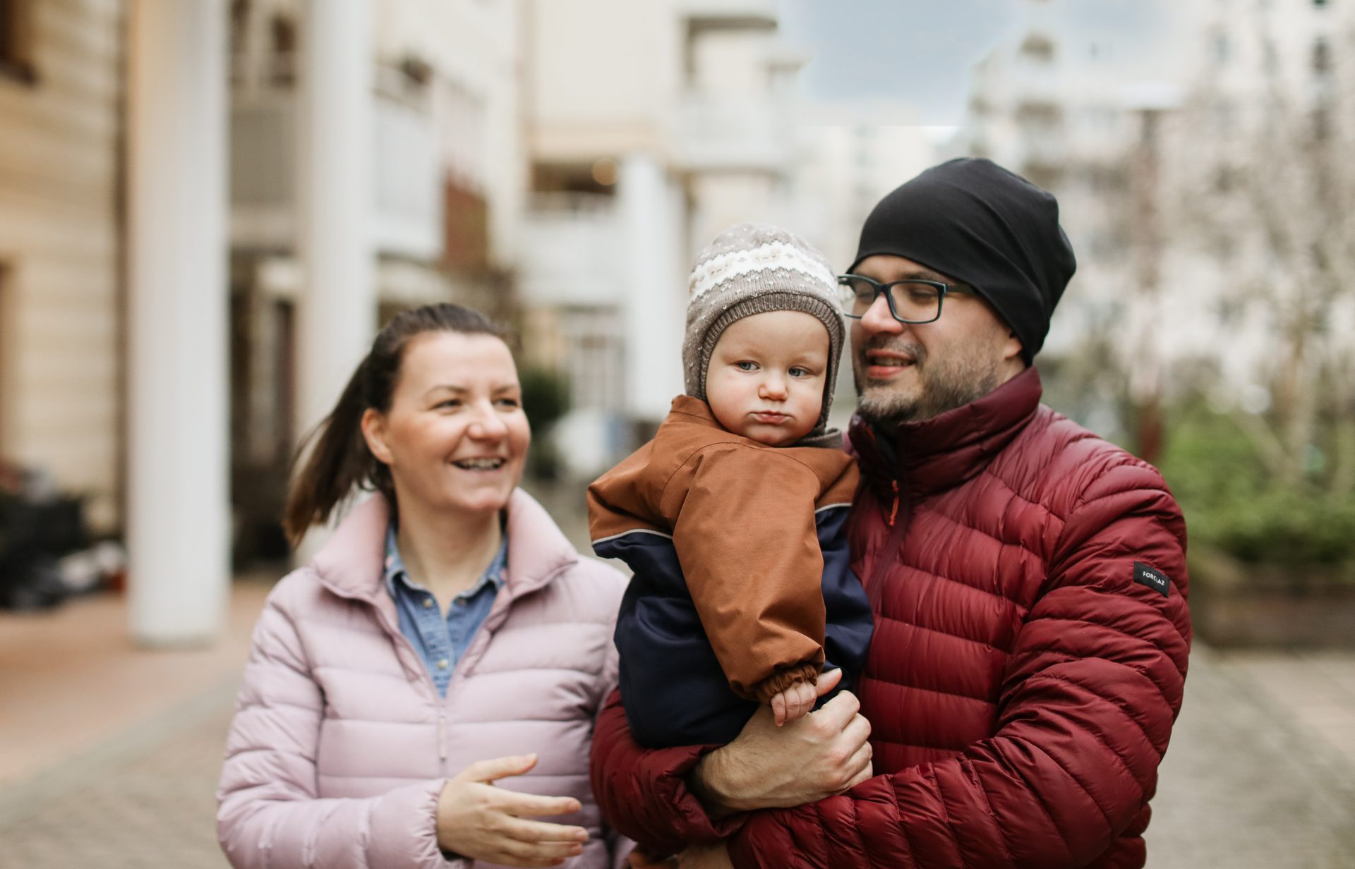 A father holds his baby in a striped hat, with a mother smiling beside them outdoors.