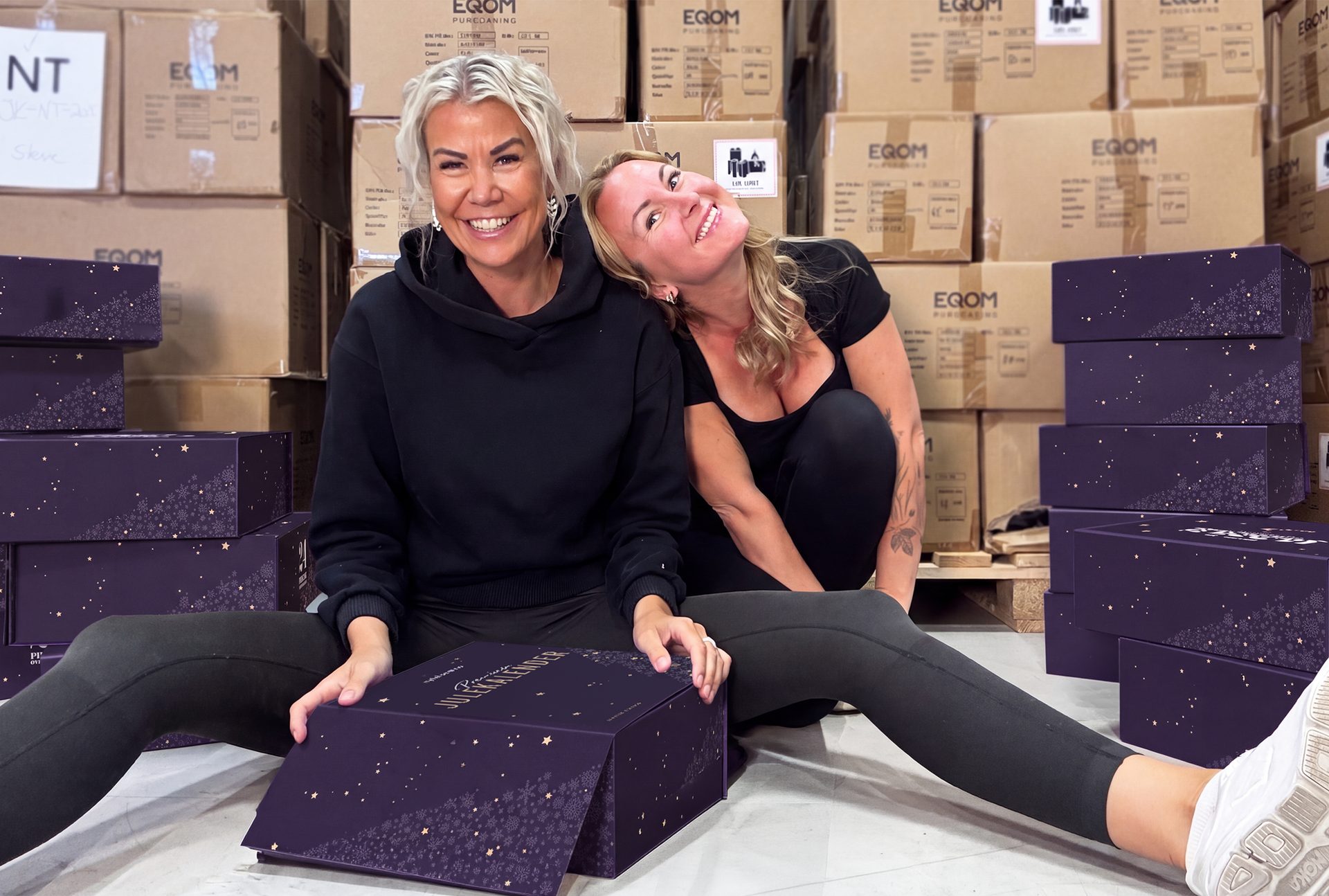 Two smiling women surrounded by many purple Christmas gift boxes and cardboard boxes in a warehouse.