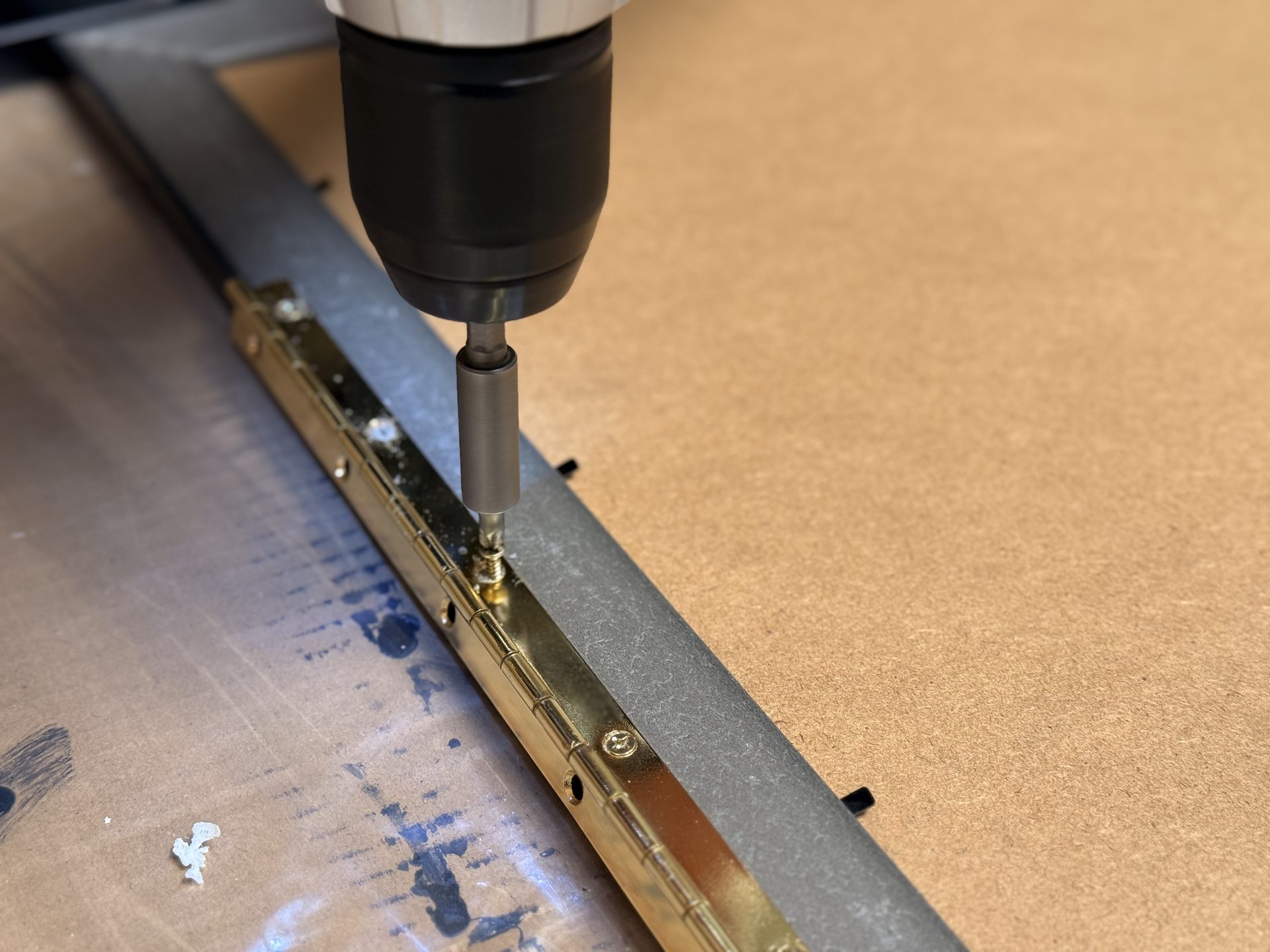 A drill drives a screw into a gold hinge attached to a wooden surface.