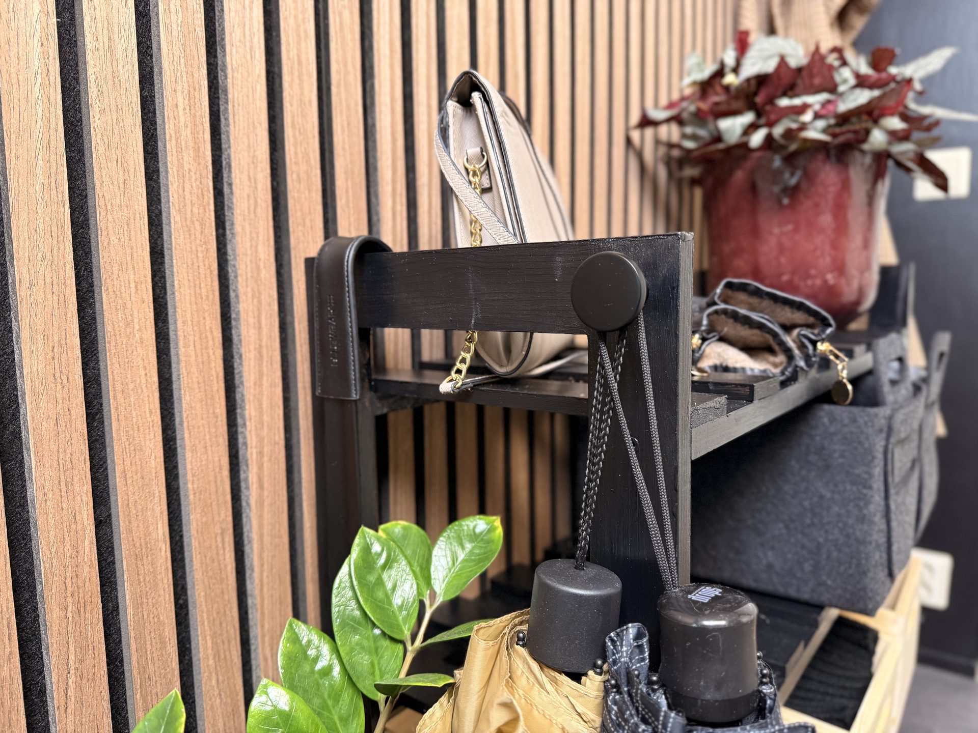 Black wooden shelf with a beige purse, green plant leaves, and umbrellas against a wood-slat wall.