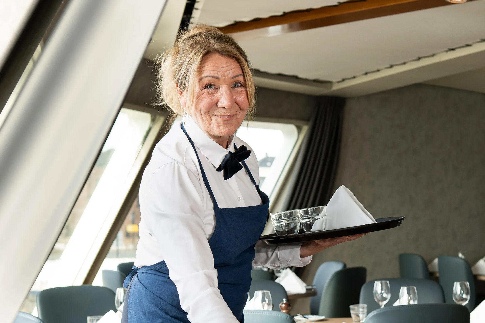 Smiling waitress in white shirt and blue apron holding a tray with glasses in a restaurant.