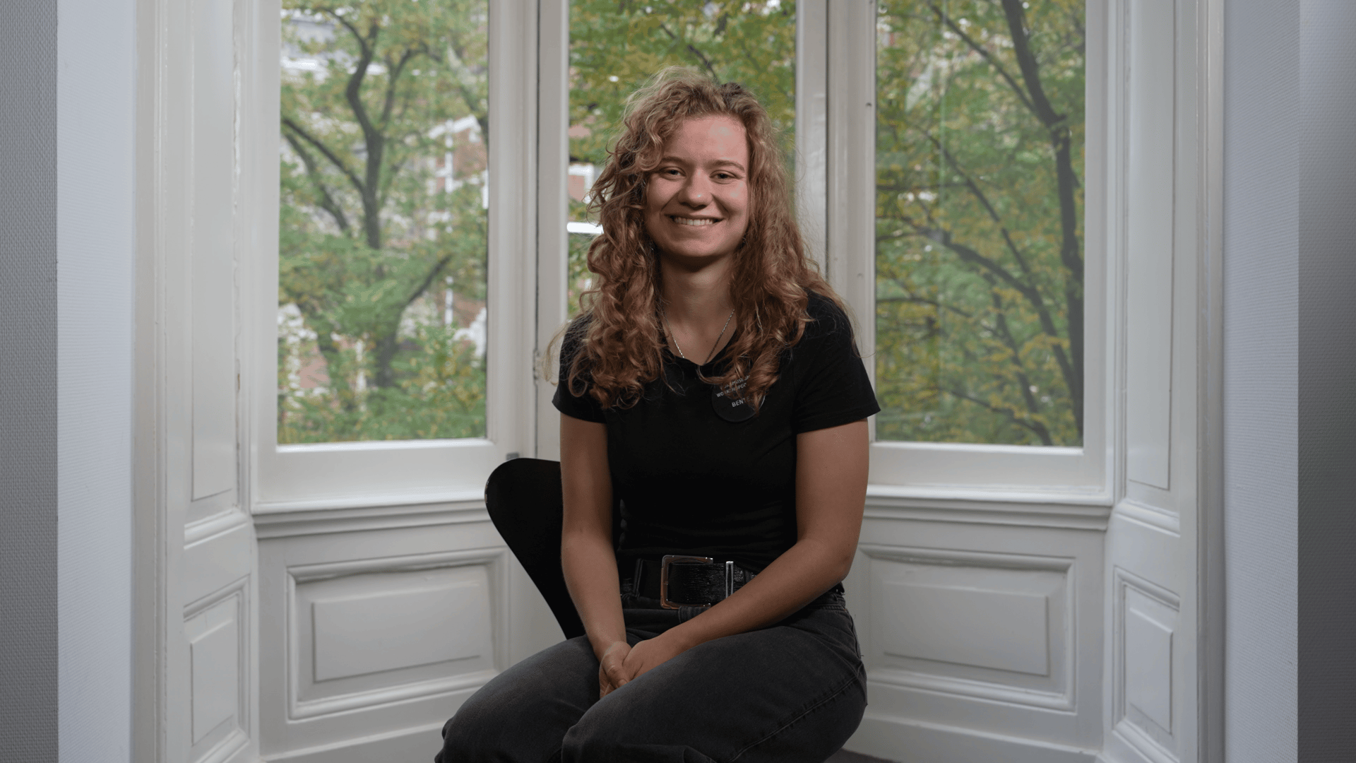 Smiling woman with curly hair, sitting by a window overlooking green trees.