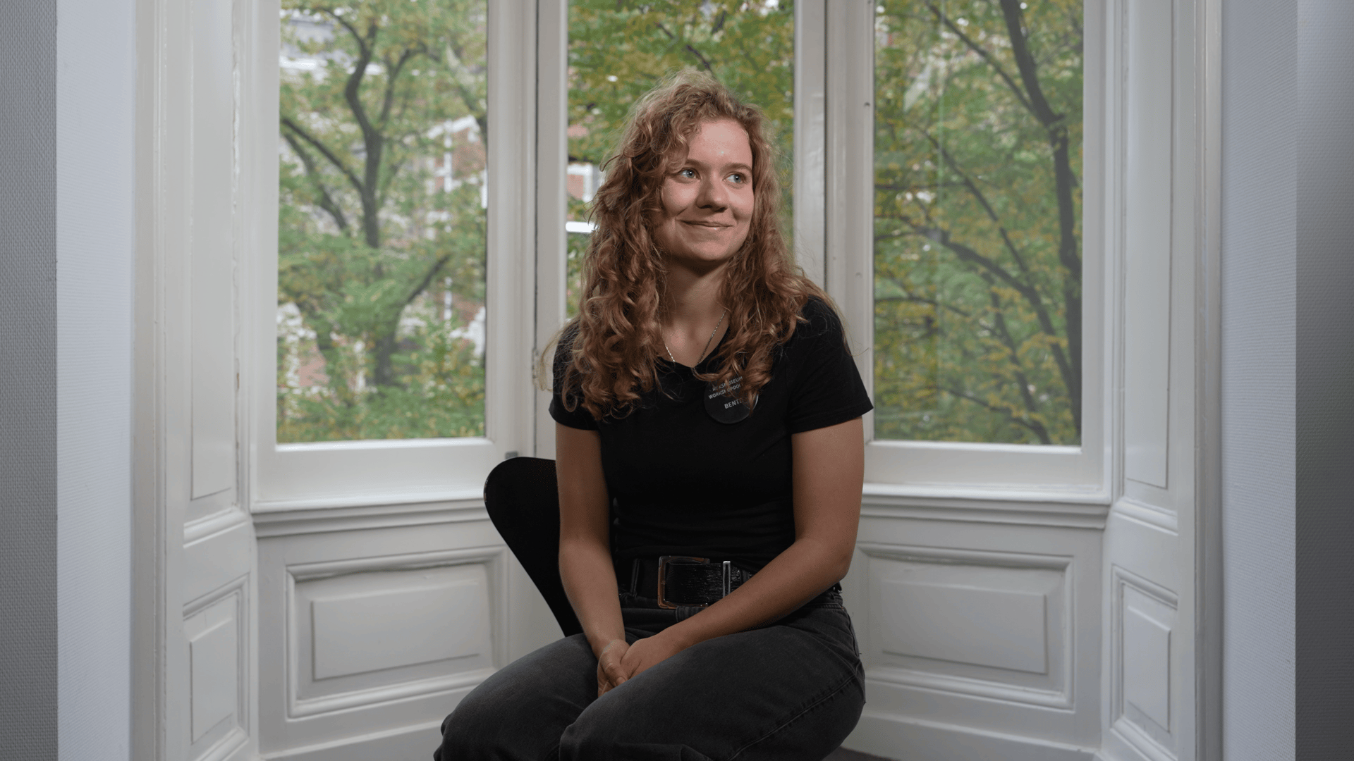 Young woman with curly hair sits in front of a window overlooking trees, smiling softly.