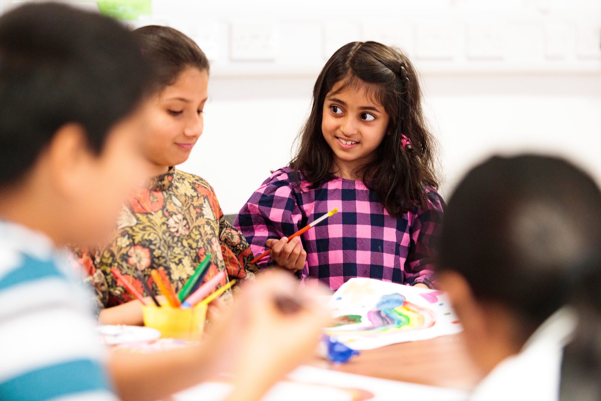 Smiling girl in pink plaid shirt painting with a paintbrush among other children.