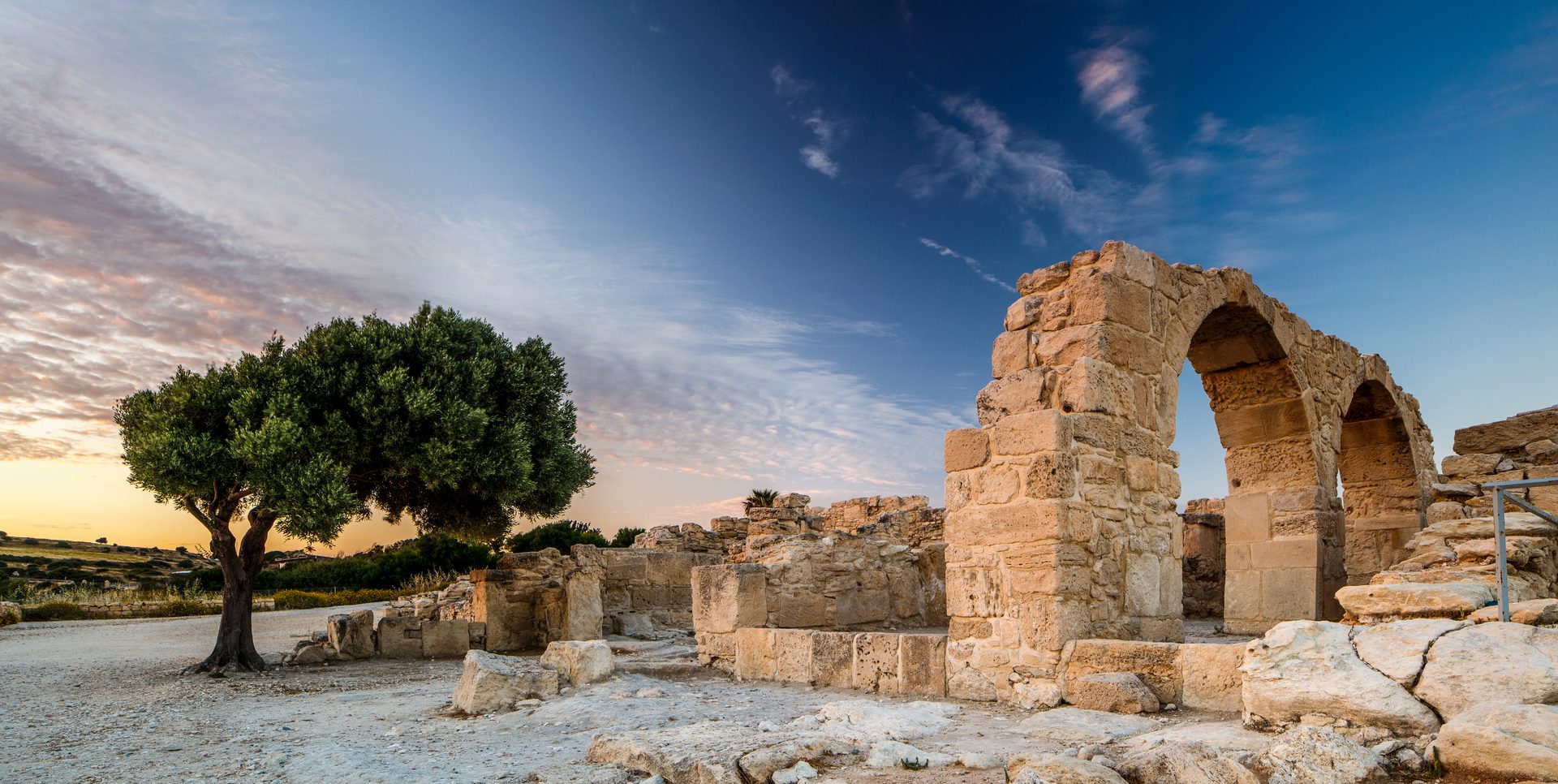 Ancient ruins with stone arches and a large olive tree under a dramatic sky at sunset.
