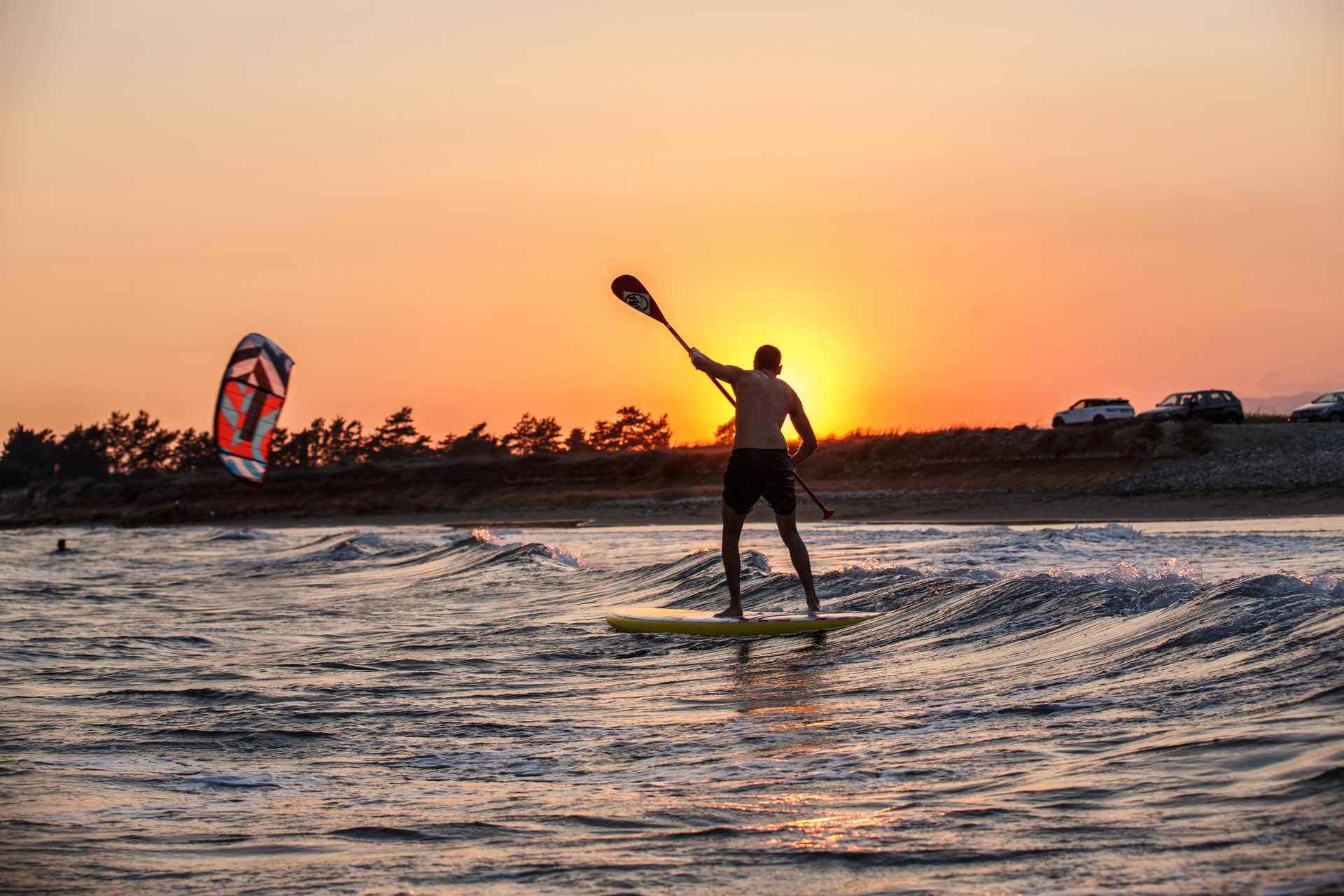 Silhouette of a man paddleboarding on ocean waves at sunset, with a kite in the distance.