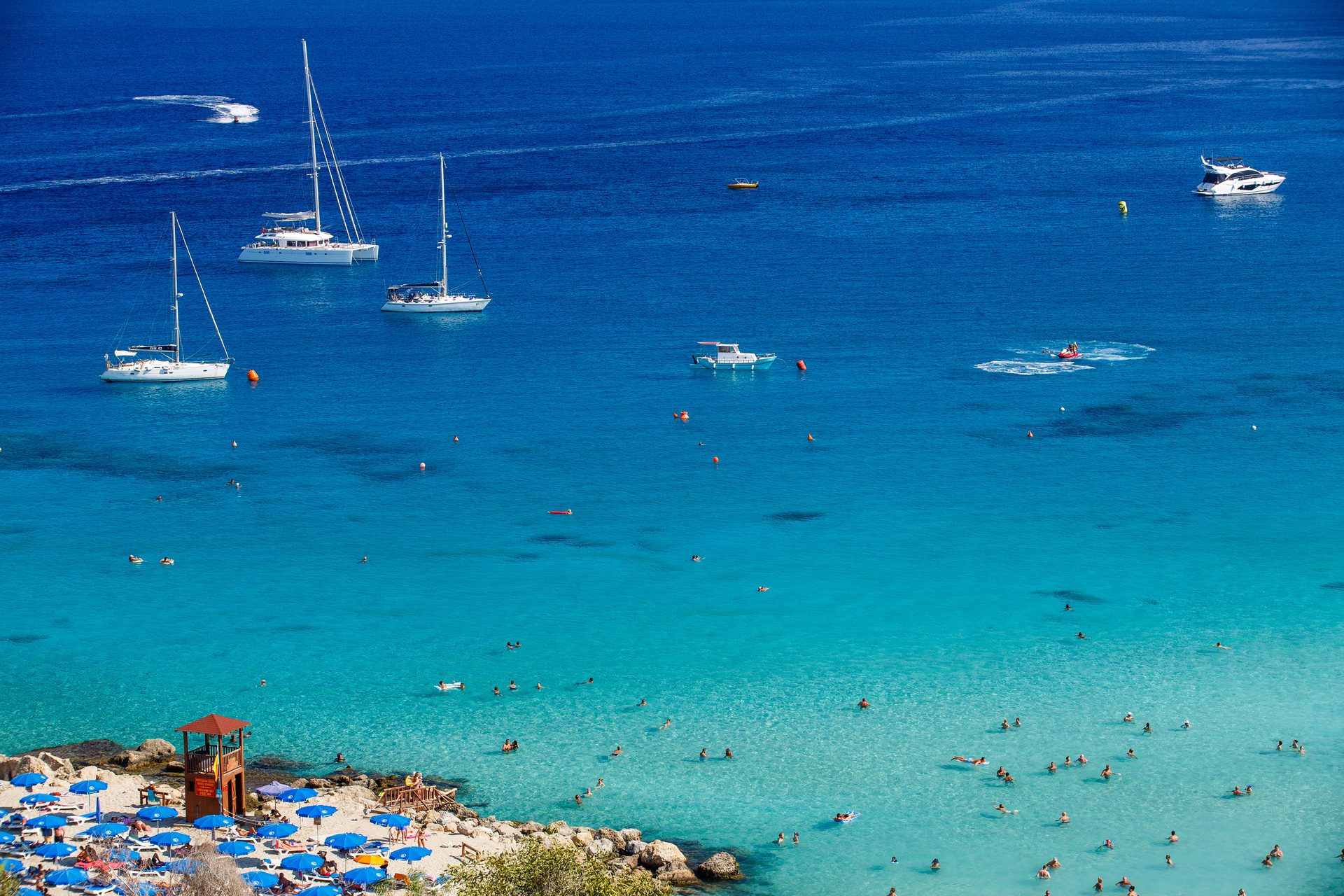 A scenic aerial view of a busy bay with boats on blue water and people swimming near a beach.