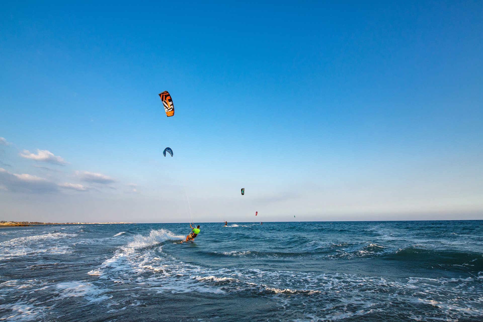 Kitesurfer riding a wave with an orange kite against a clear blue sky, other kites in distance.