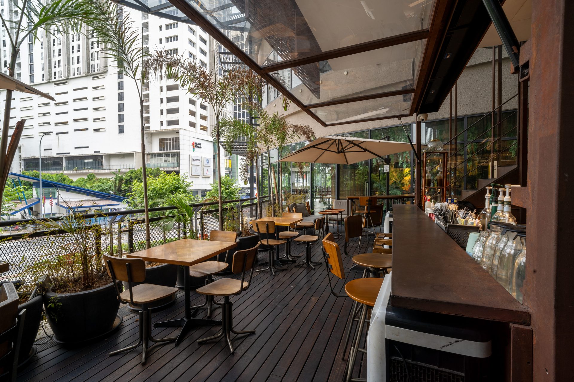 Outdoor cafe patio with wooden tables, chairs, and a bar, overlooking city buildings.