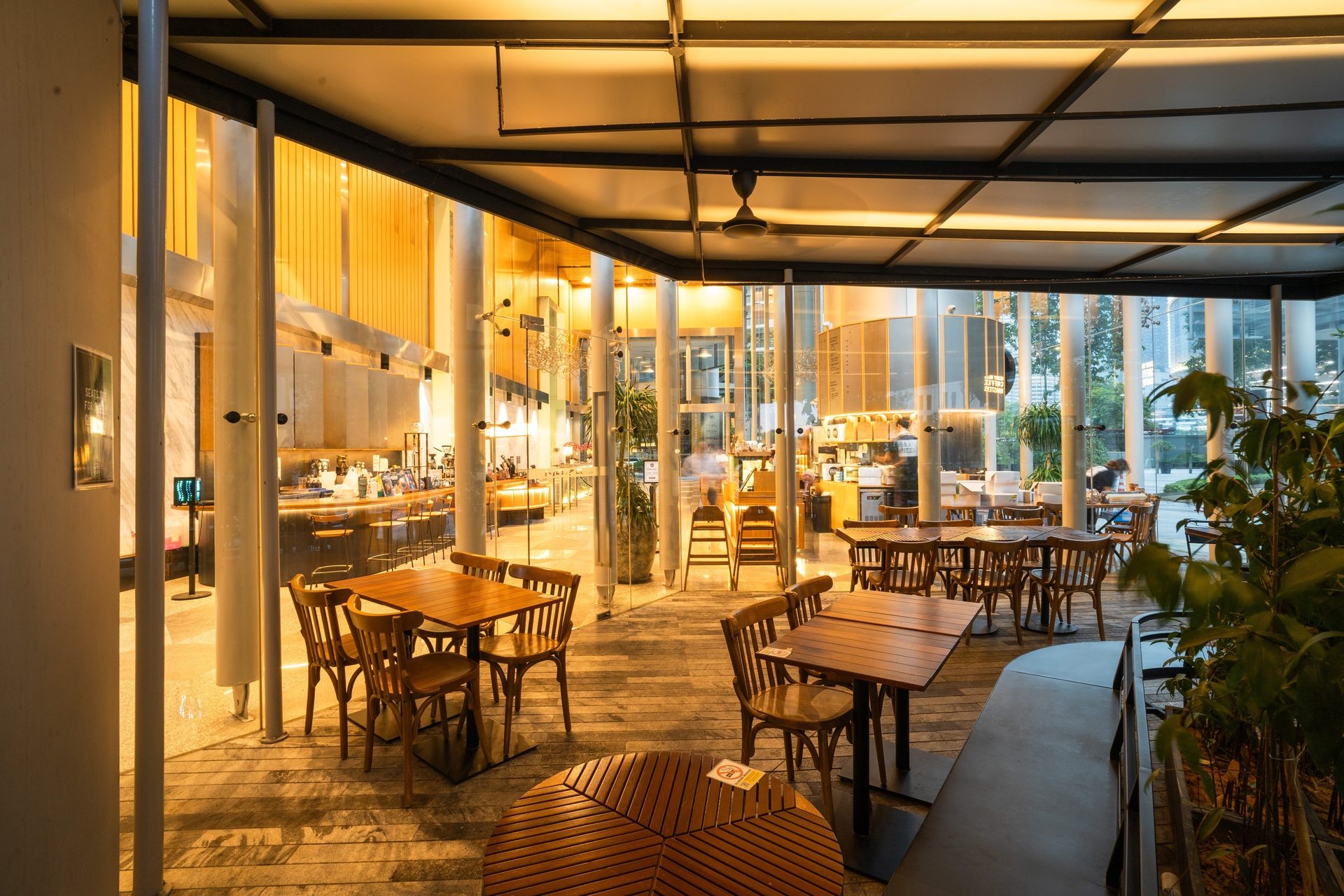 Warmly lit modern cafe interior with wooden tables, bar, and large windows.