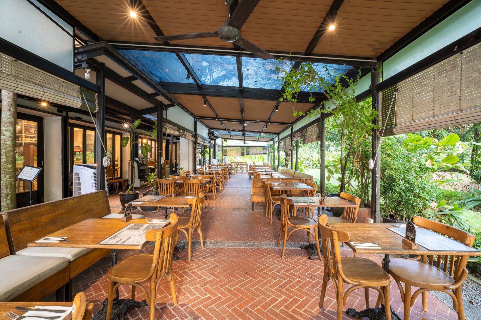 Patio restaurant with rows of wooden tables on a brick floor, next to lush outdoor greenery.