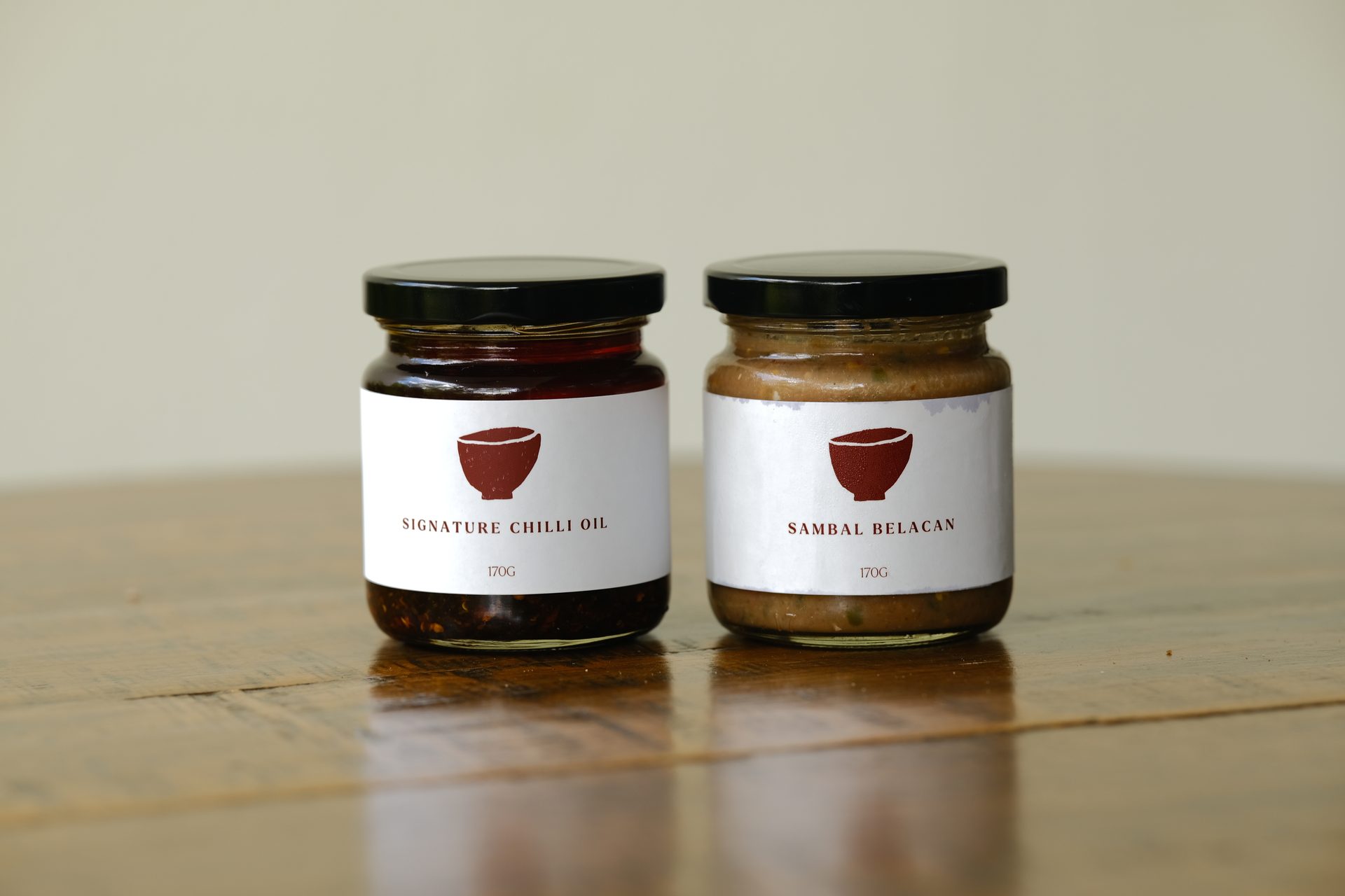 Two jars of chili oil and sambal belacan with red bowl icons on a wooden table.