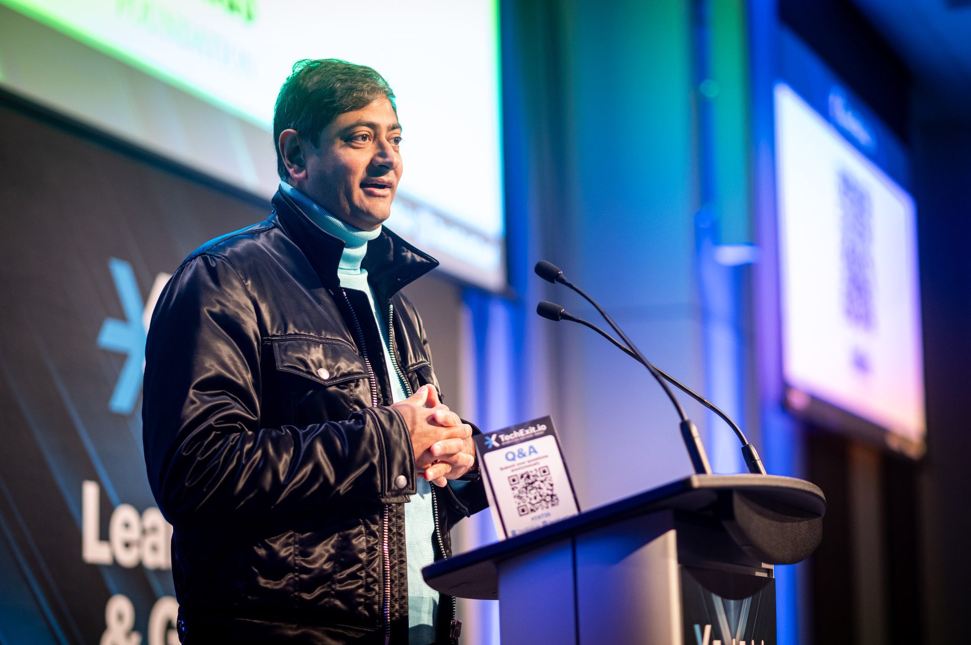 A man in a black jacket and blue turtleneck speaking at a conference podium with microphones and a Q&A sign.