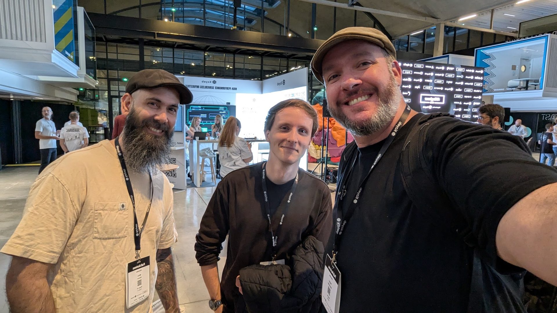 Selfie of three smiling men with lanyards at an indoor event with exhibition booths.