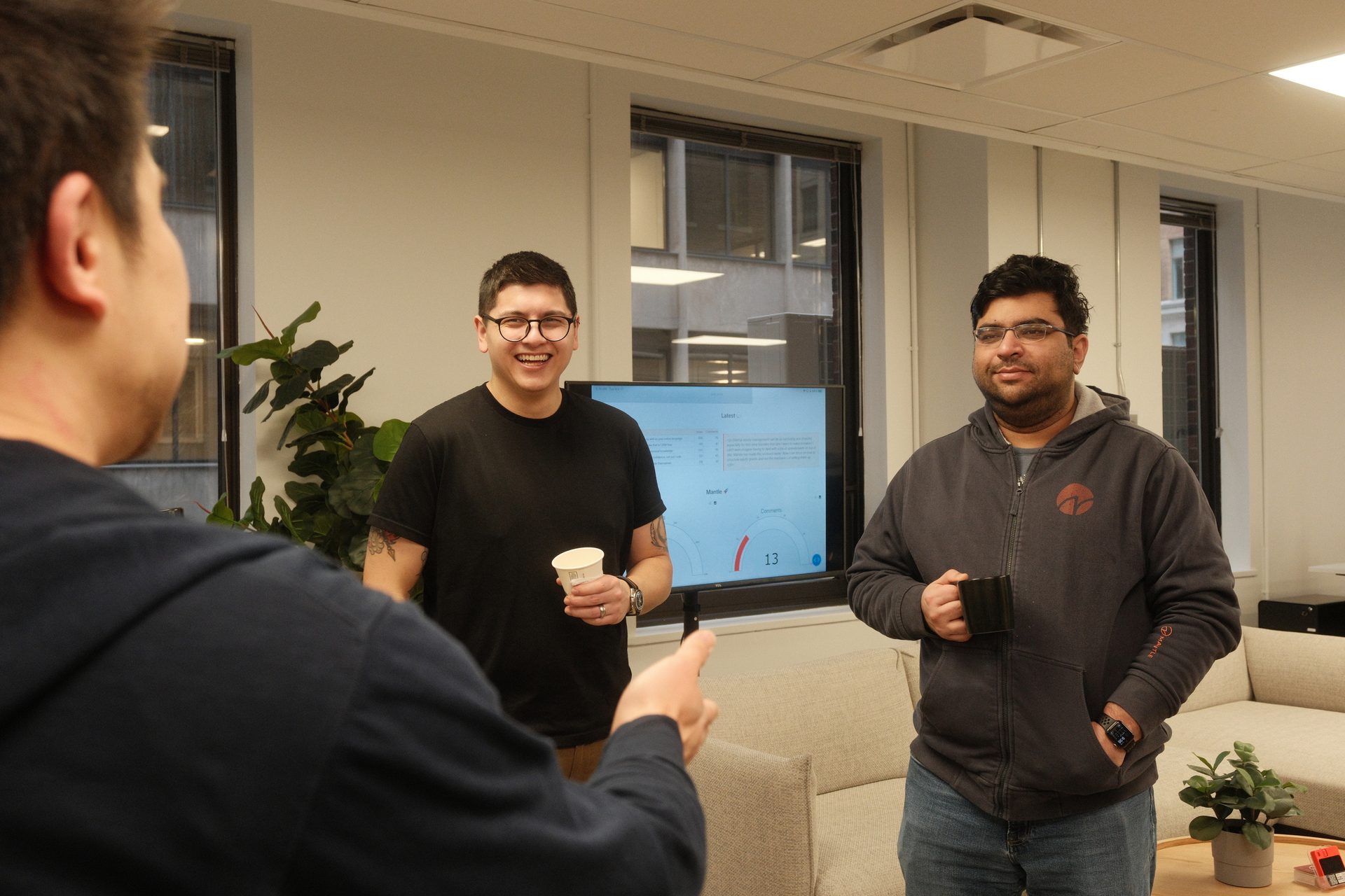 Three men discussing in an office, with a data screen in the background.