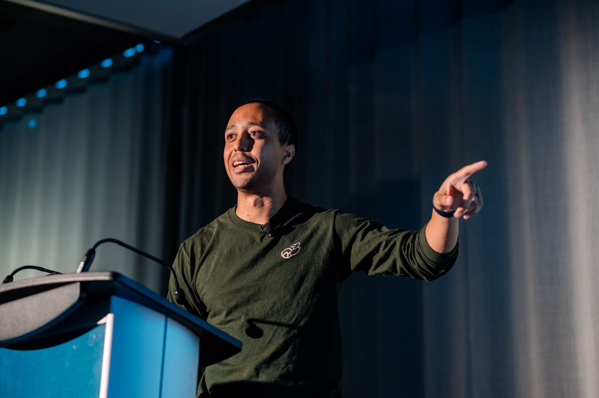 A man in a dark green shirt speaks animatedly at a podium, pointing to the right.