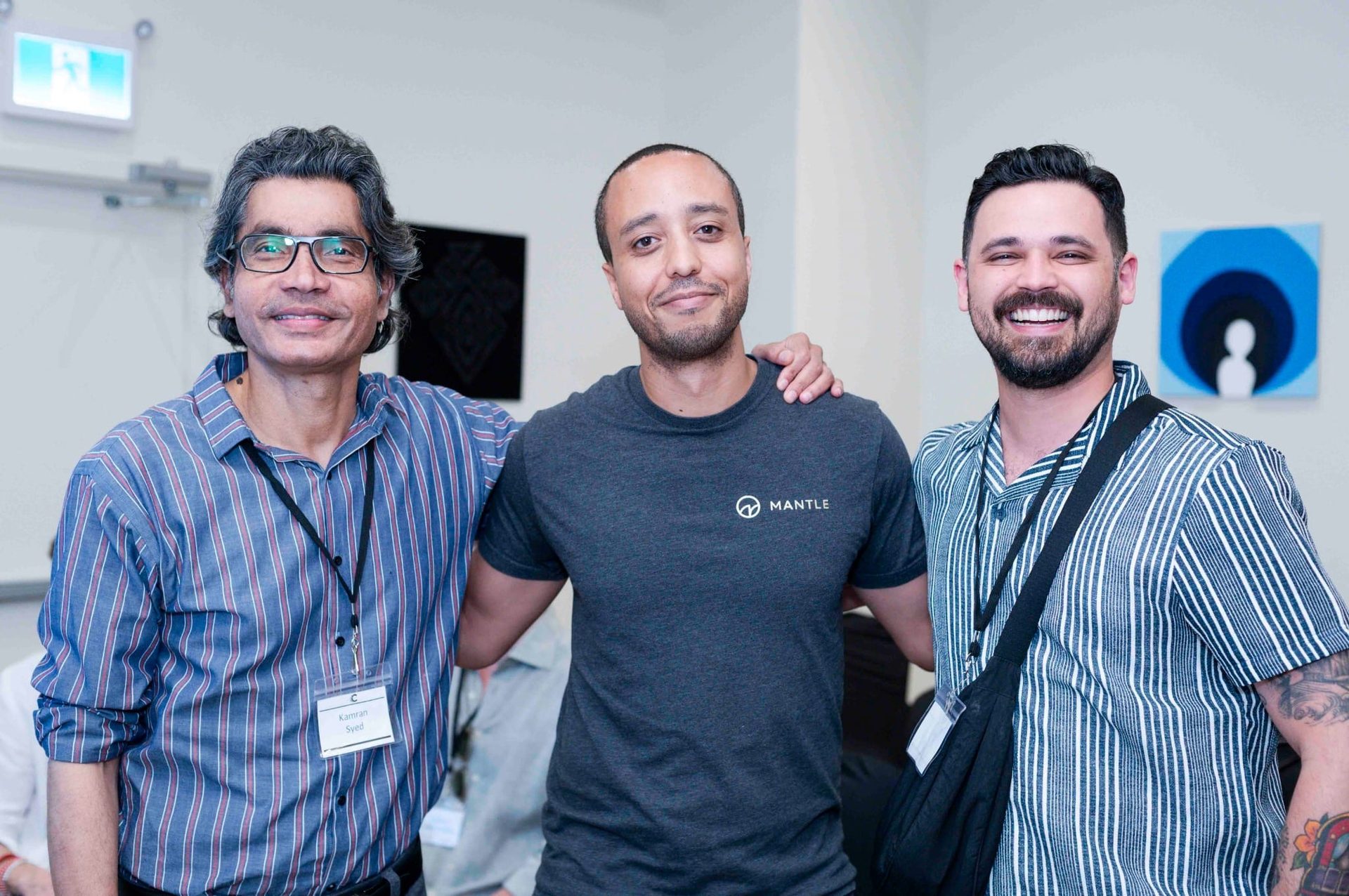 Three smiling men; one wears a Mantle shirt, another has a "Kamran Syed" name tag.