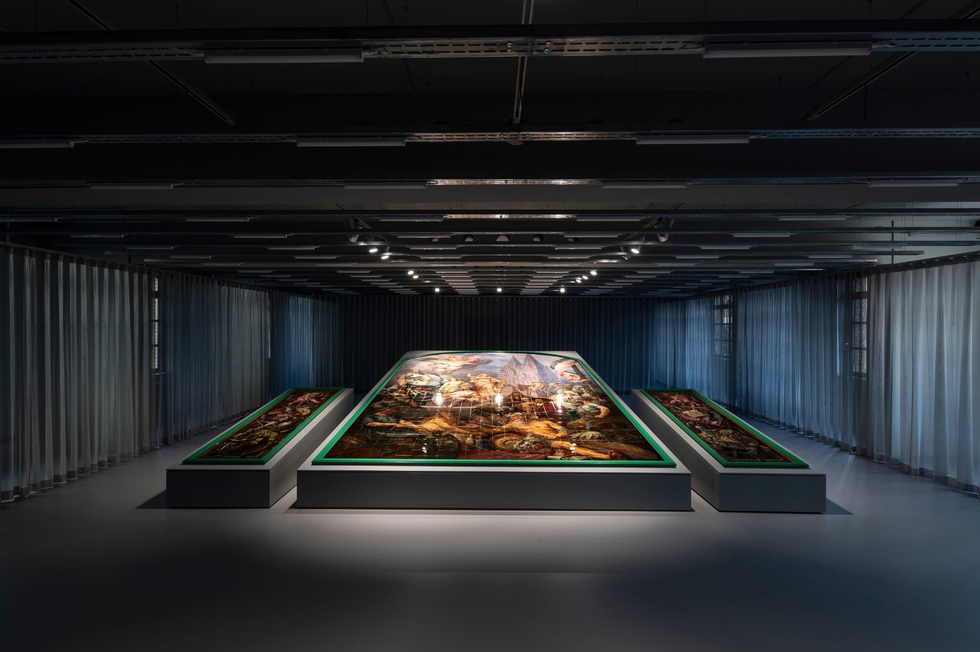 Three large, green-framed art displays on platforms in a dark exhibition room with curtains.
