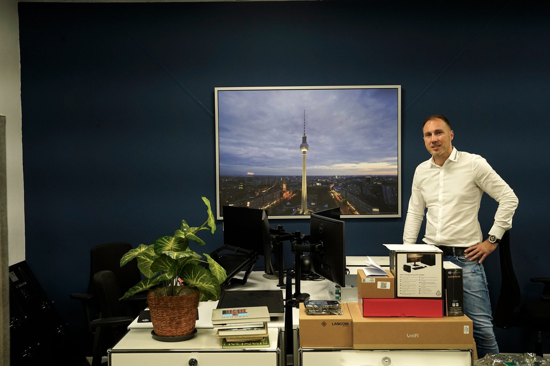 Man in an office with a Berlin TV Tower picture and boxes on a desk.