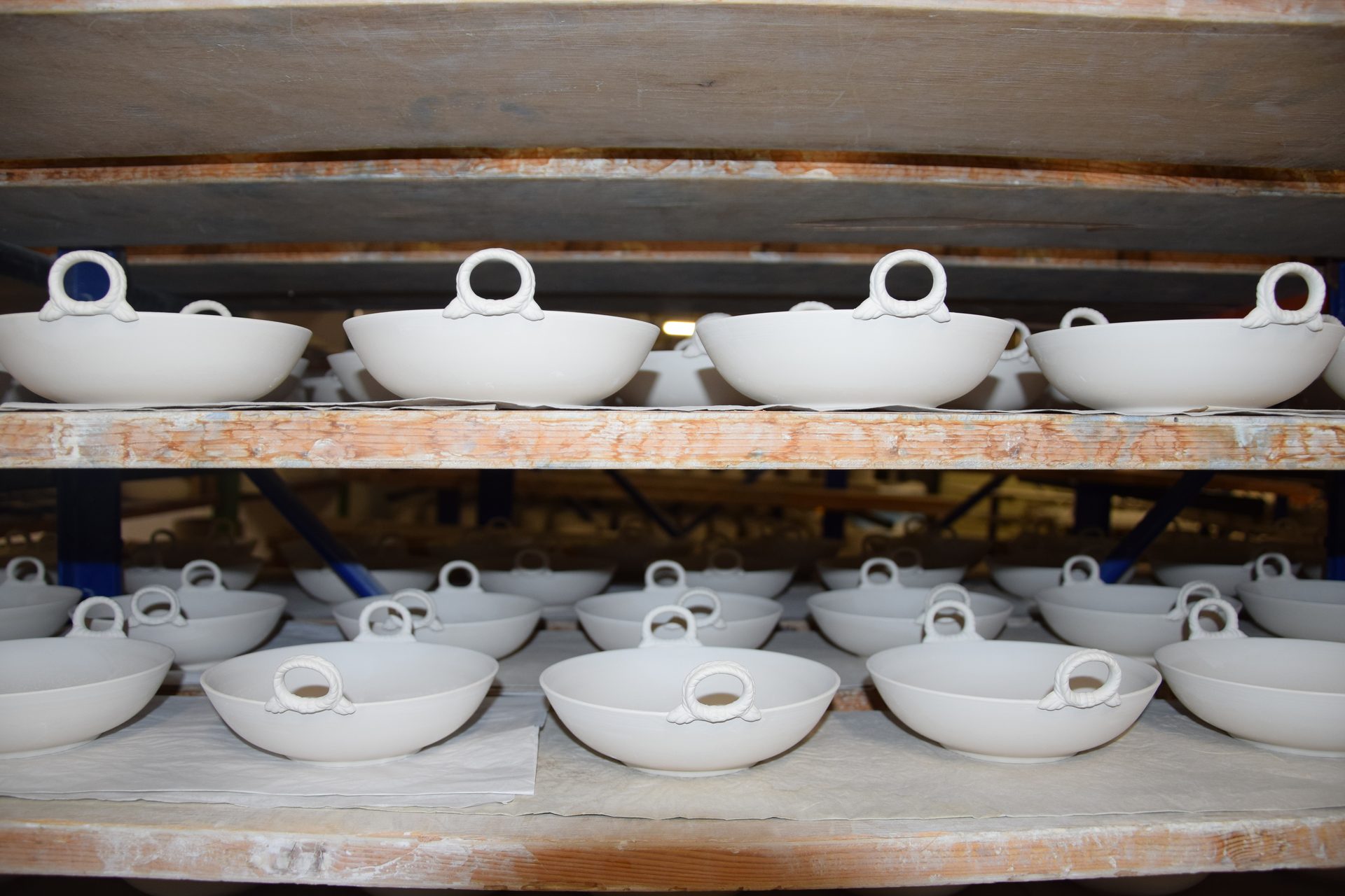 White ceramic bowls with handles drying on wooden shelves in a pottery studio.