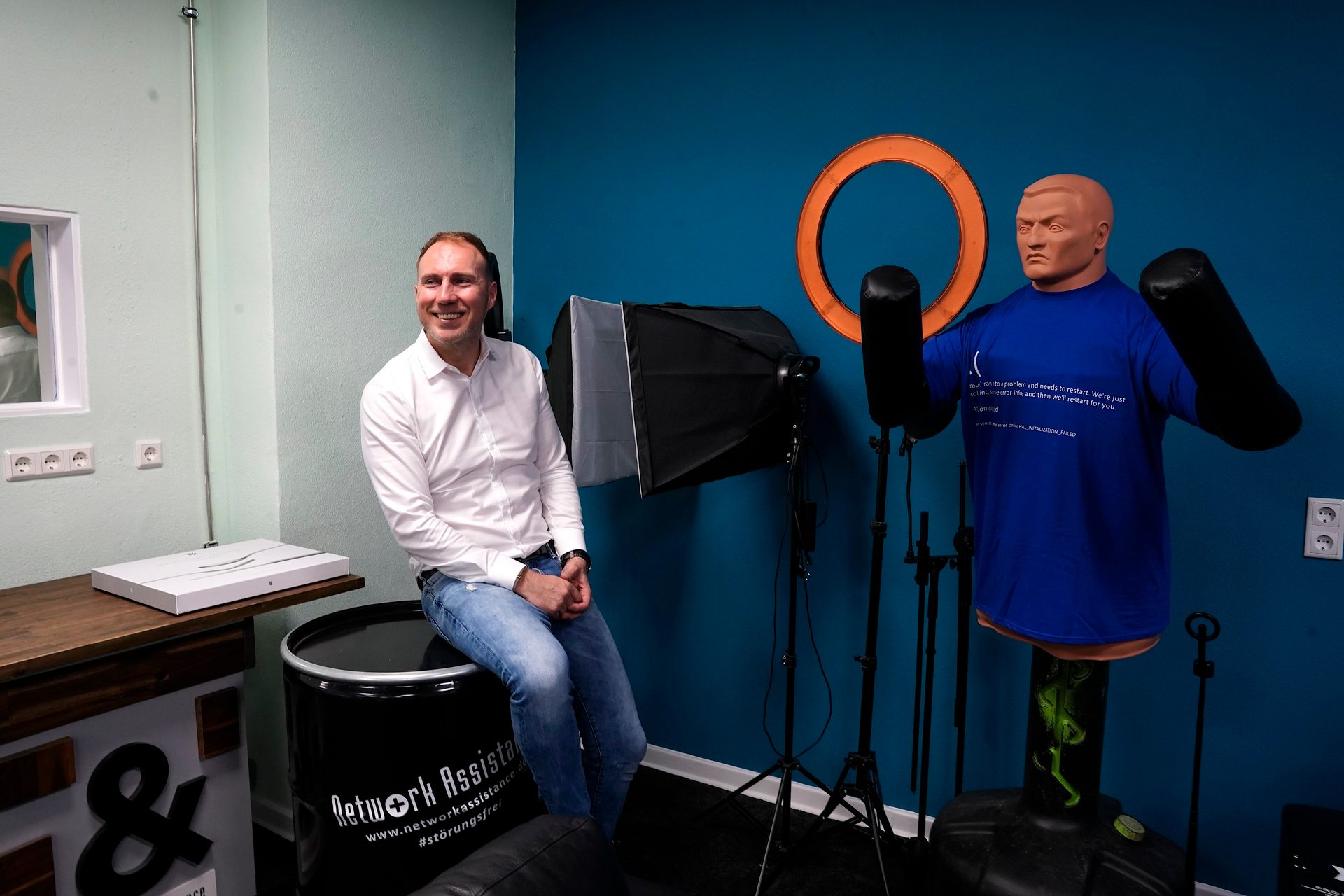 A smiling man sits in a studio with a punching dummy, lighting equipment, and "Network Assistance" branding.