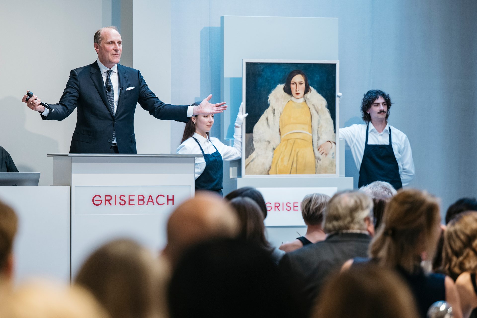 Auctioneer at Grisebach presents a painting of a woman in yellow and fur, flanked by two assistants.