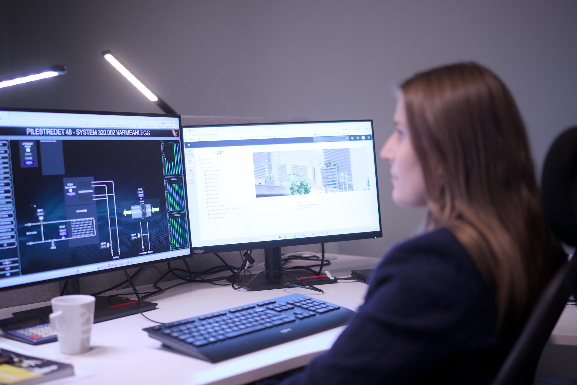 Woman working at a desk with two computer monitors displaying data and a website.