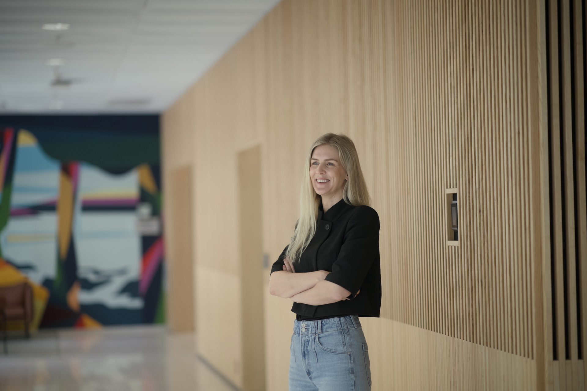 A smiling blonde woman in a black jacket and jeans stands with arms crossed in a modern hallway.